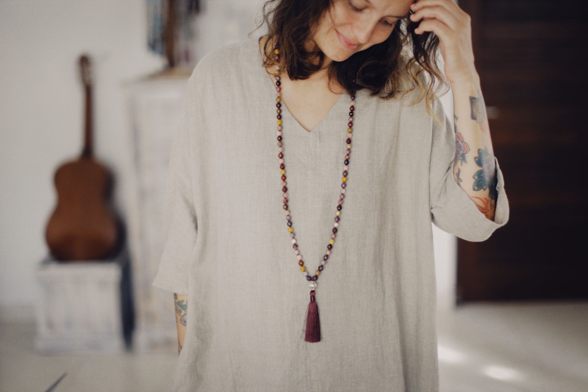 A woman with tattoos wearing a long beaded necklace, standing indoors with soft lighting.