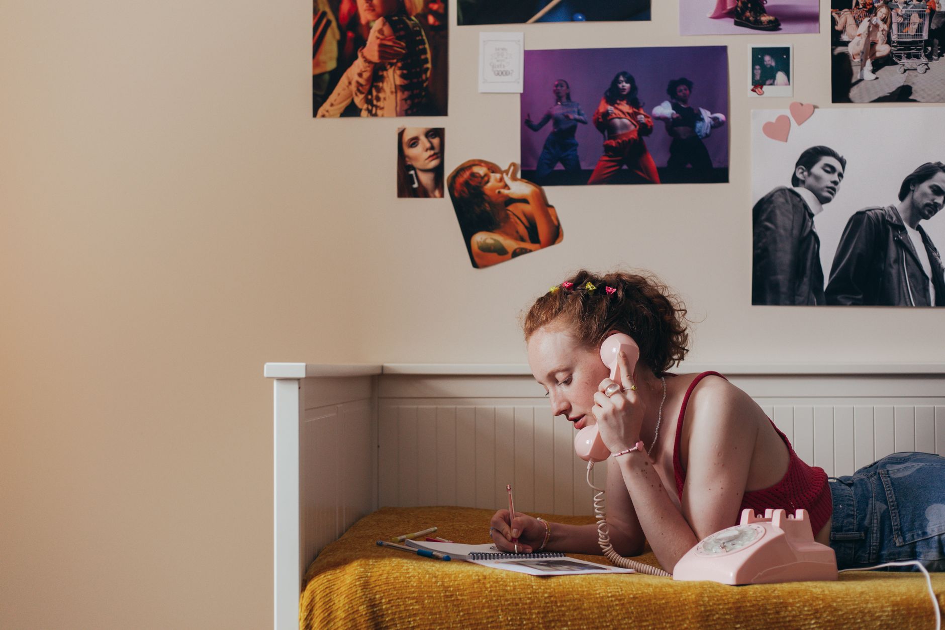 Teen girl writing notes on bed while talking on a retro pink telephone, 90s nostalgia.
