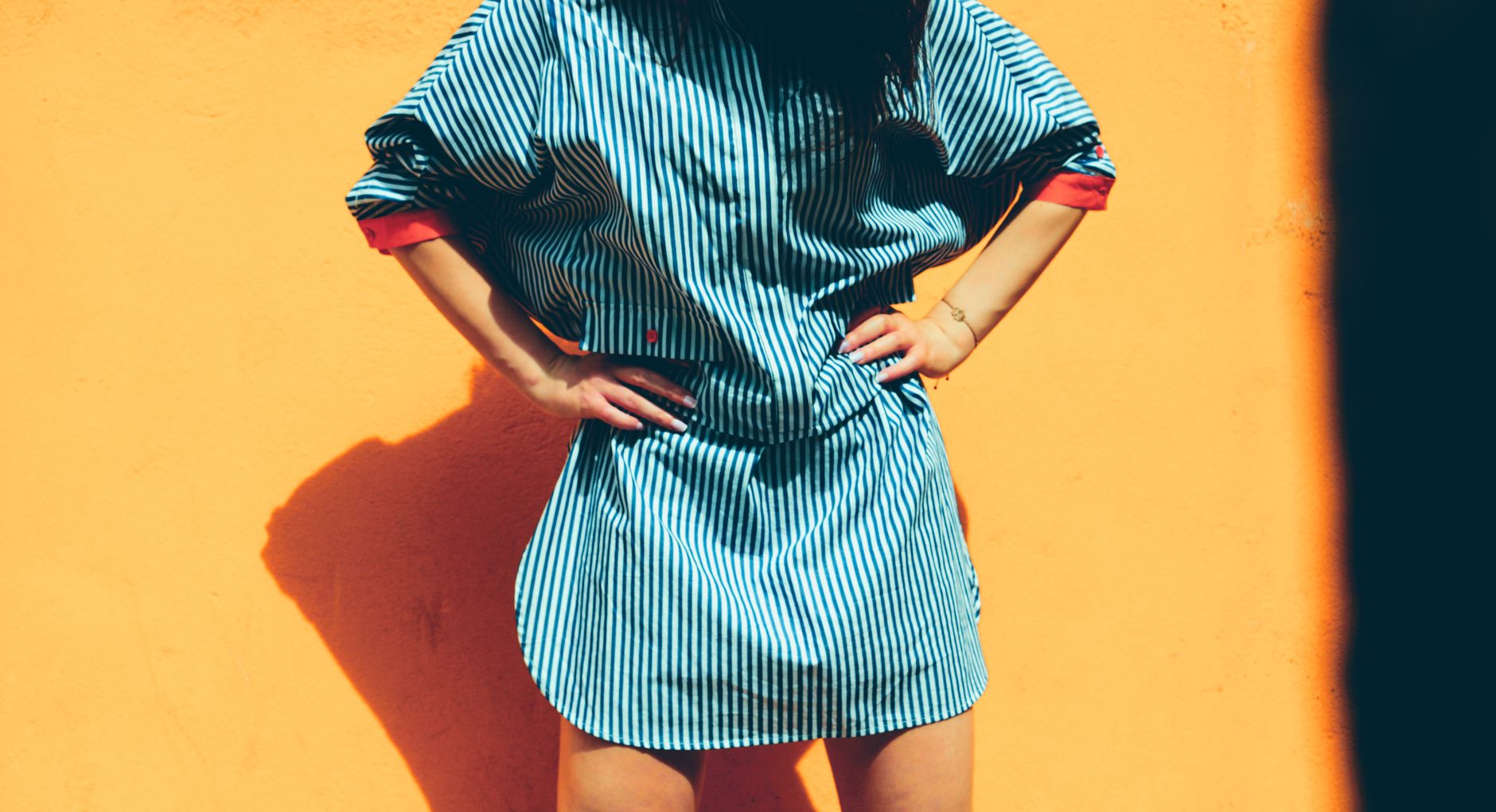 Fashionable woman posing in striped dress against a vibrant orange wall in sunny light.