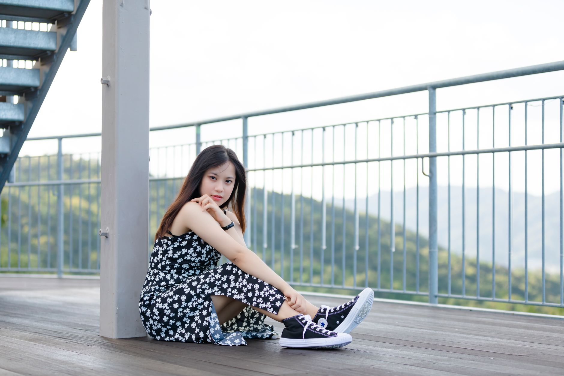 Asian woman in floral dress sitting outdoors, posing on a balcony with a serene view.