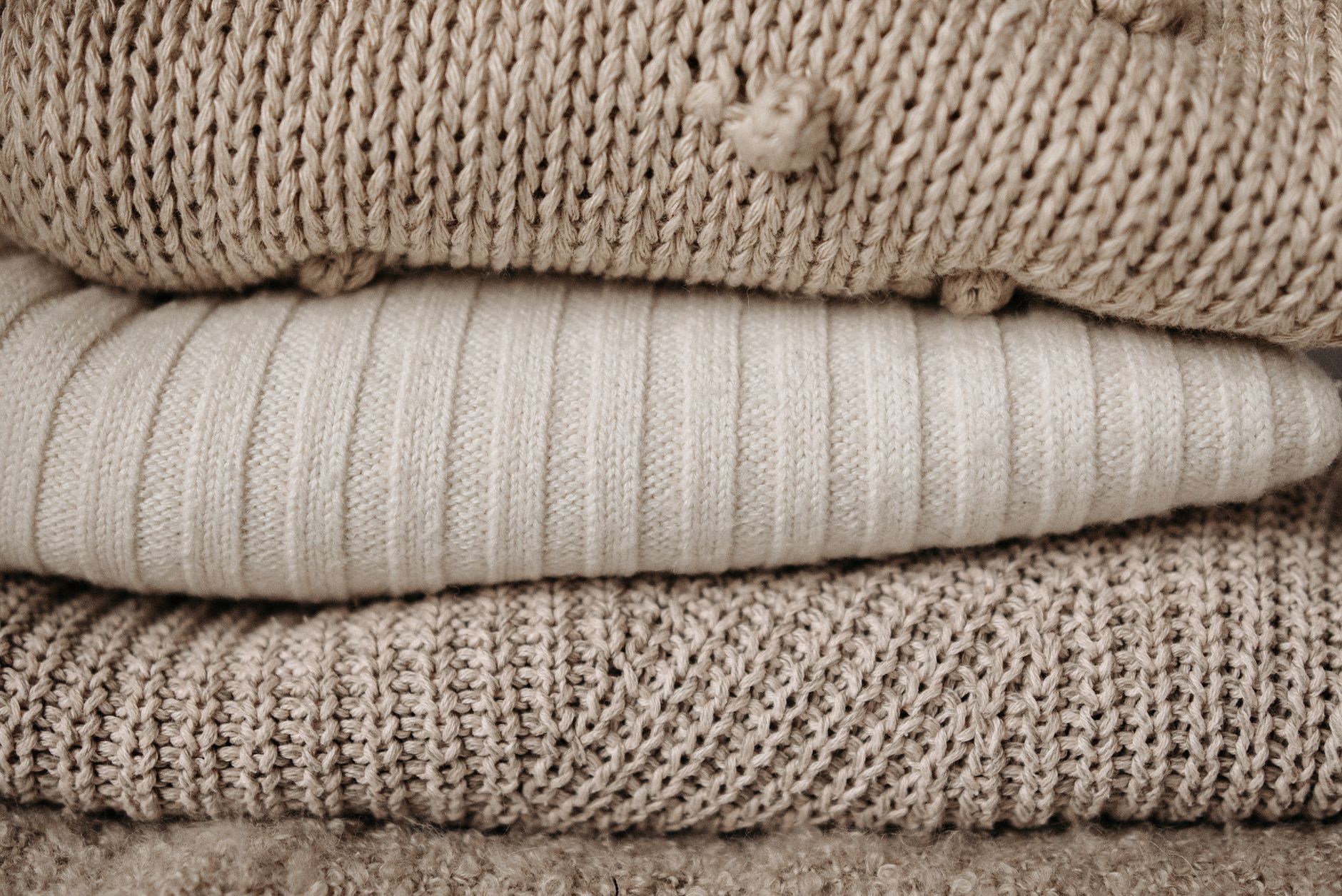 Textured close-up of stacked beige knit sweaters showcasing various knitting patterns.