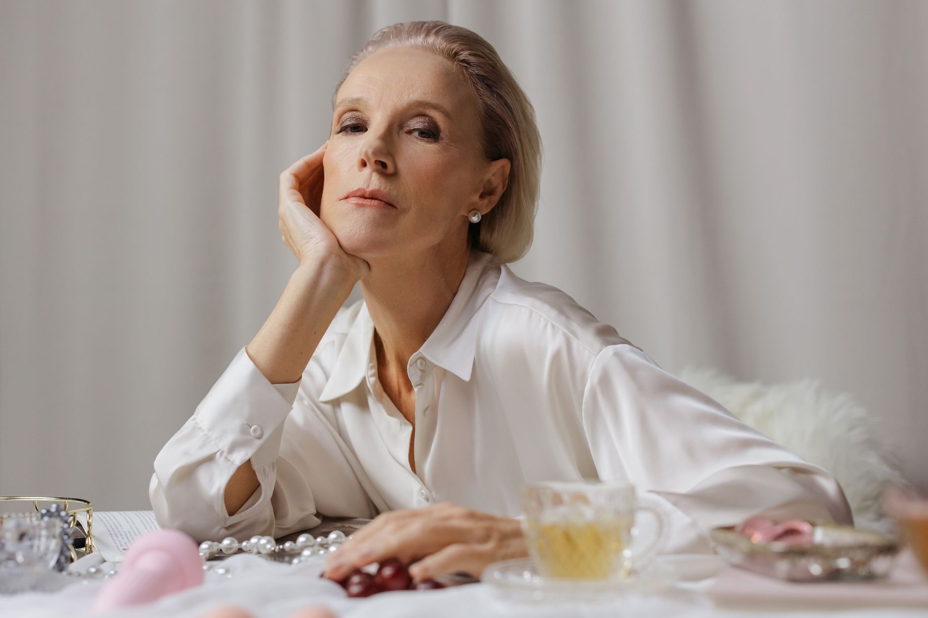 A poised senior woman in a white shirt enjoying tea, surrounded by elegant pearls.