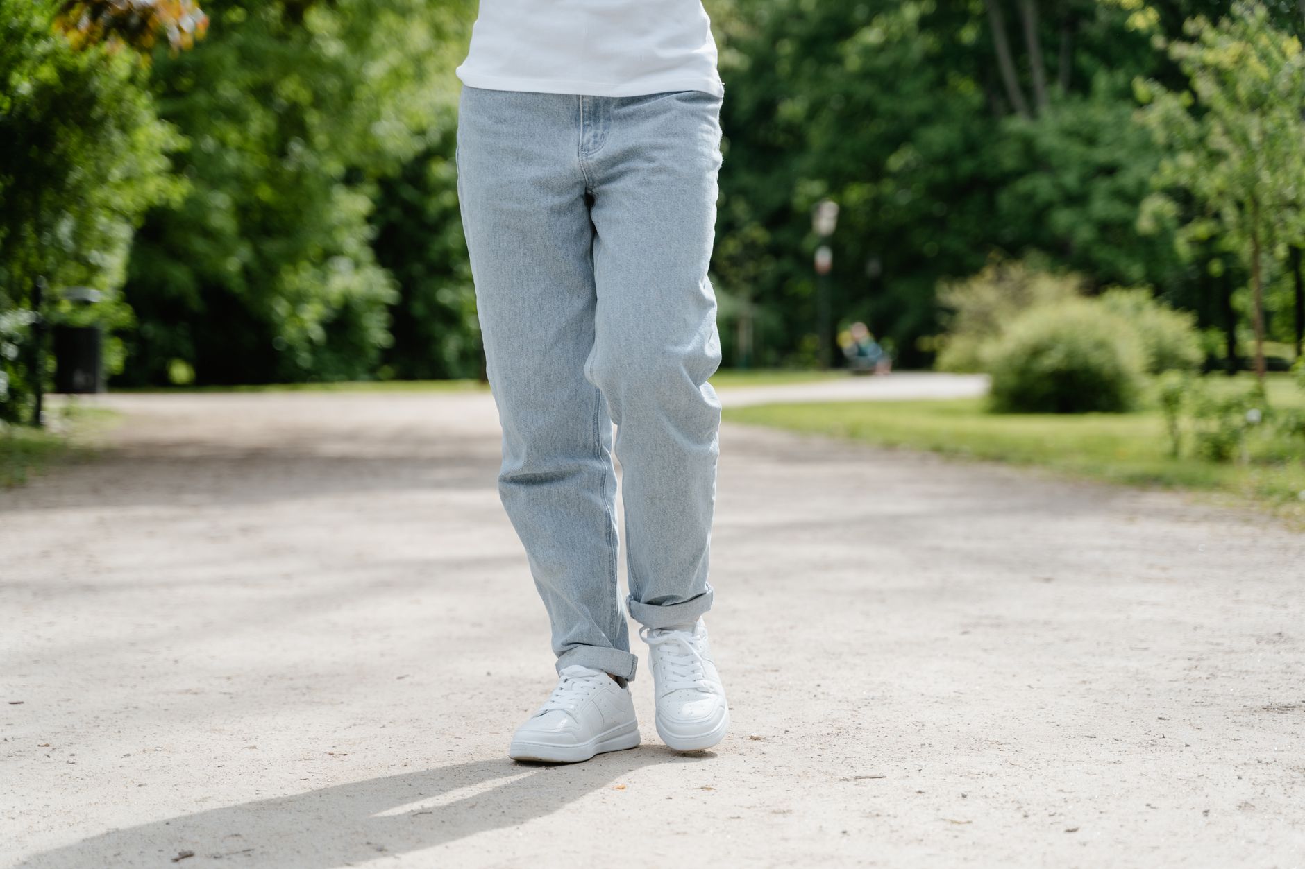 A close-up view of a person walking on a park pathway wearing blue jeans and white sneakers.
