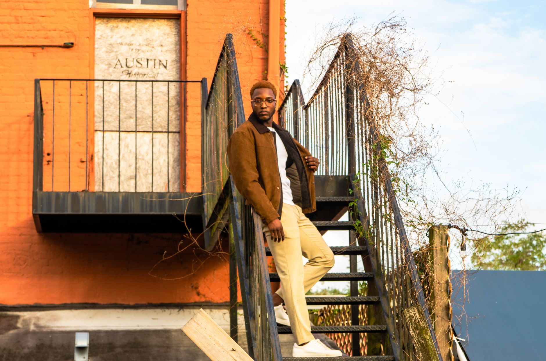 Stylish man in suede jacket posing on staircase beside an orange building.