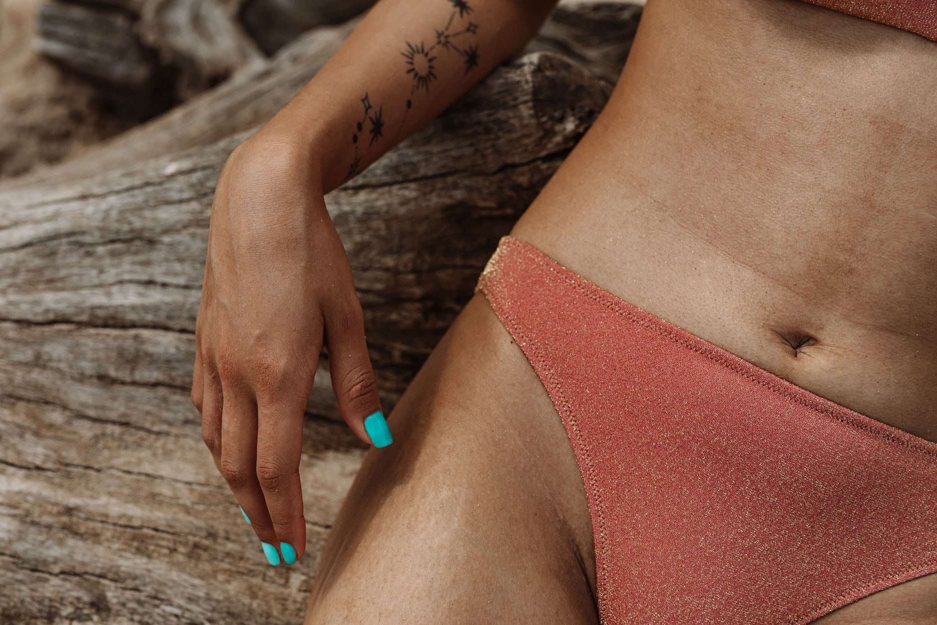 A close-up of a bikini-clad woman's tattooed arm and manicured nails resting on a tree trunk.