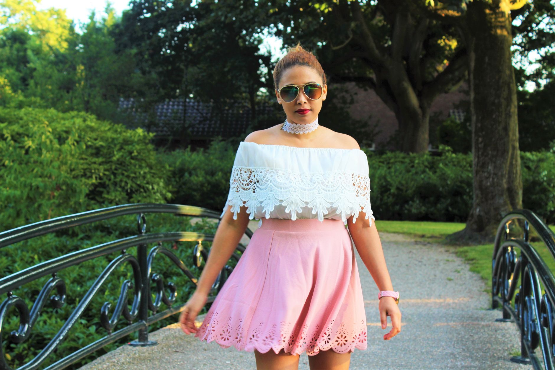 A young woman in fashionable attire poses confidently on a garden bridge under daylight.