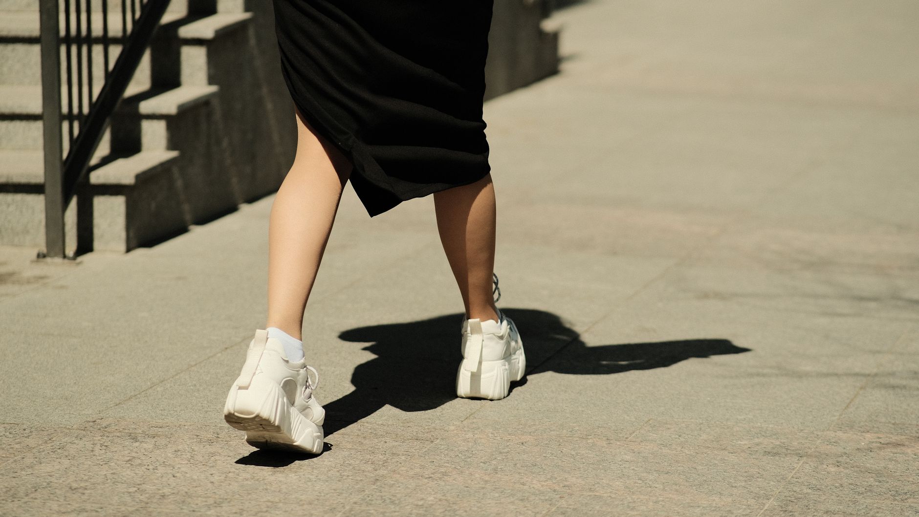 A person in a black dress walks outdoors on a sunny day wearing white sneakers.