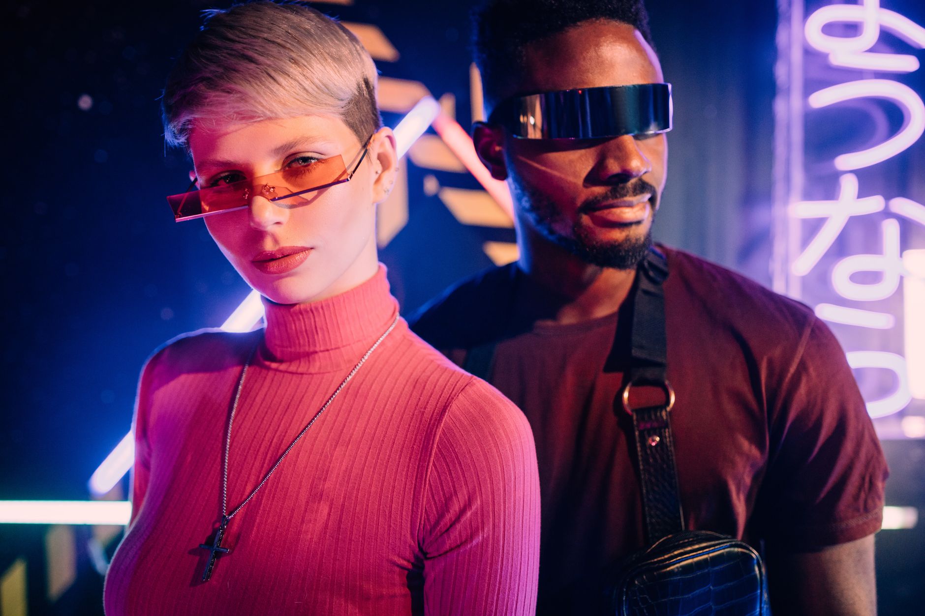 Stylish interracial couple in neon-lit setting, showcasing futuristic eyewear and fashion.