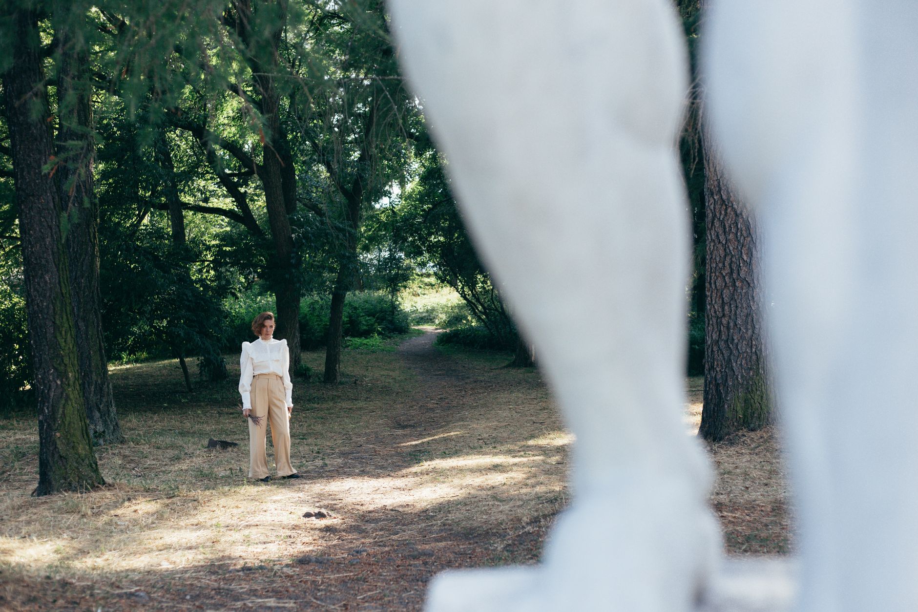 Woman in white blouse and beige pants stands beside a marble statue in a tree-lined park.