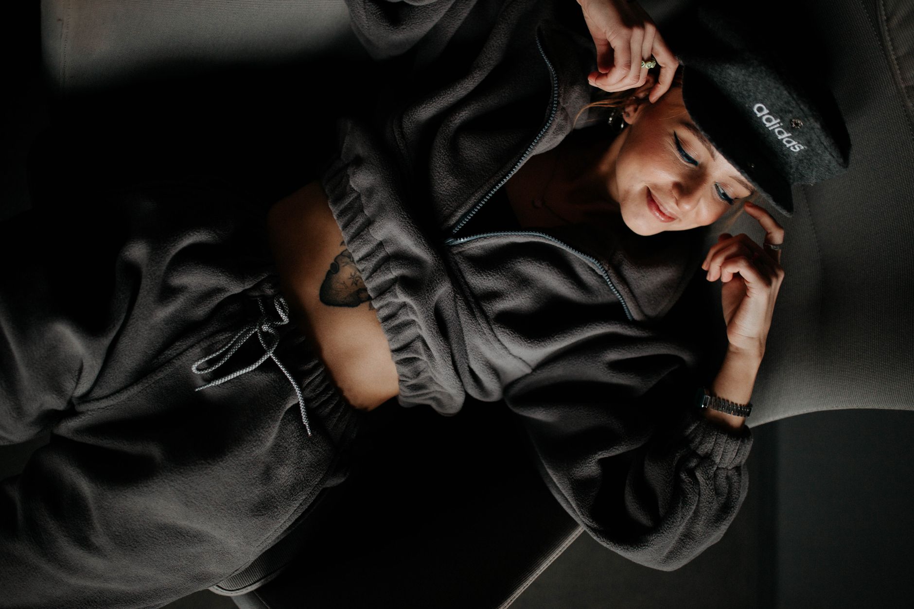 A stylish woman in an Adidas hat and loungewear poses comfortably indoors, showcasing modern fashion.