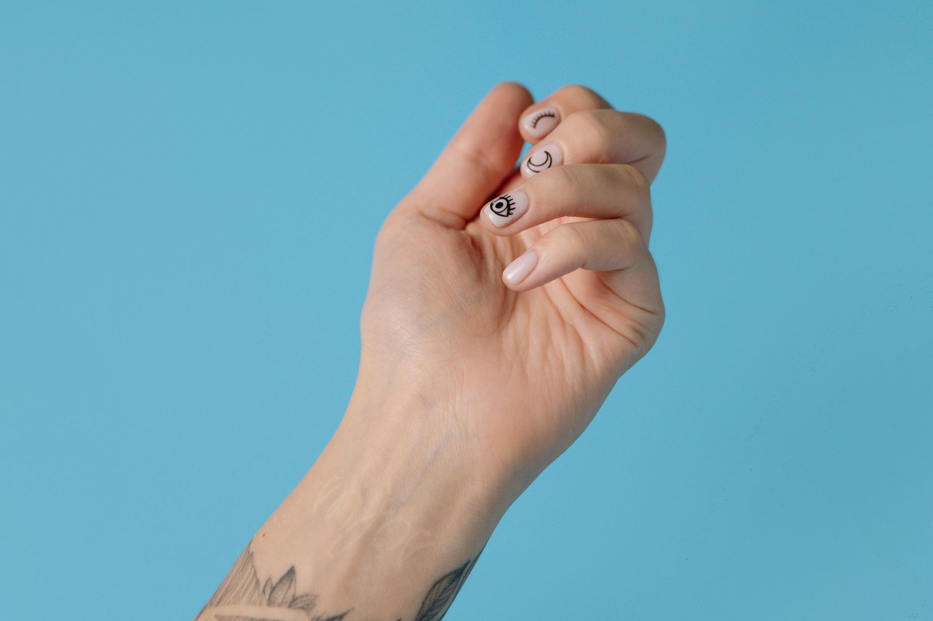 Close-up of a tattooed hand with artistic nail art against a pastel blue background, showcasing creativity and style.