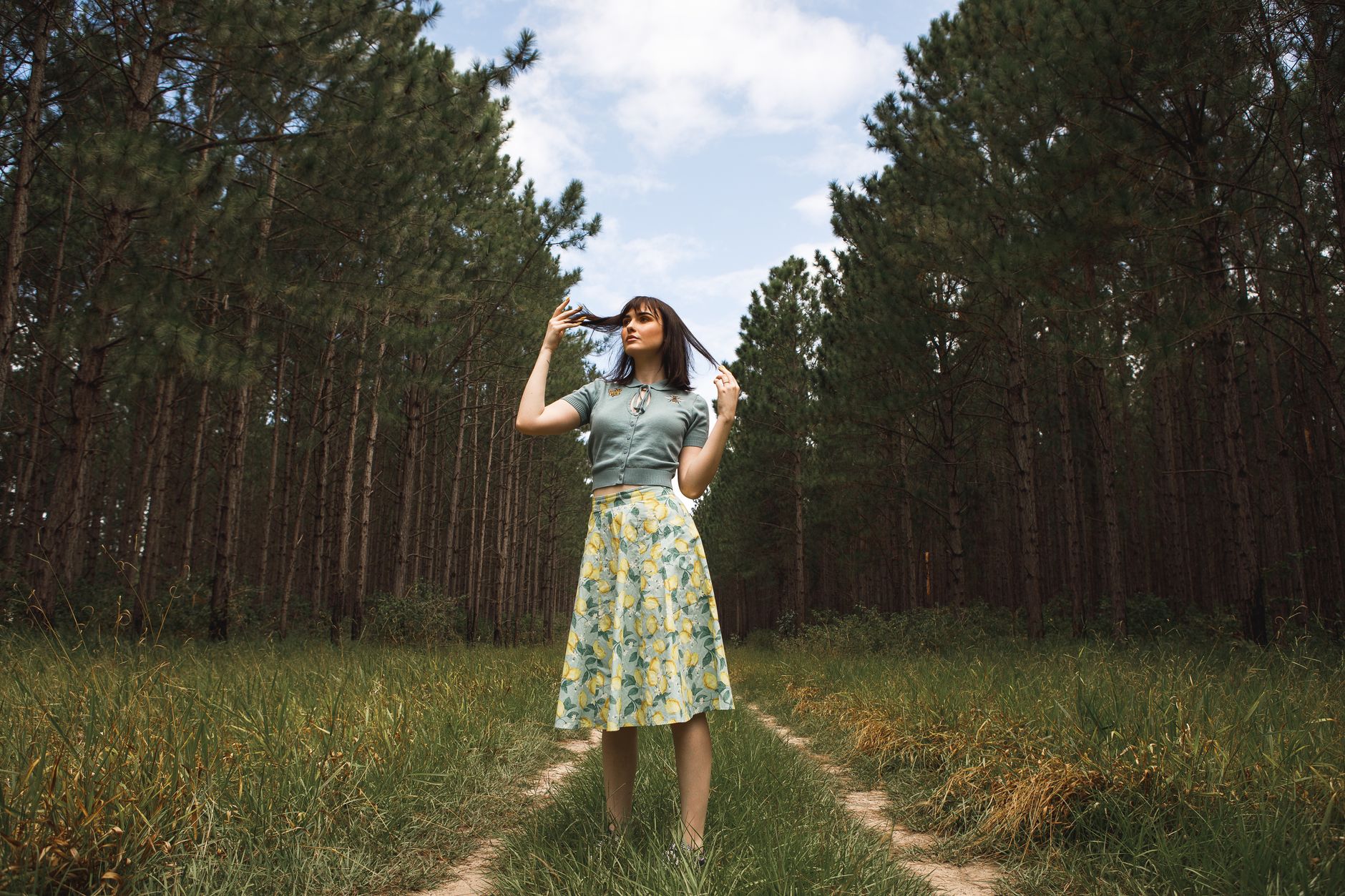 Elegant woman in a floral skirt stands on a forest path surrounded by tall trees.
