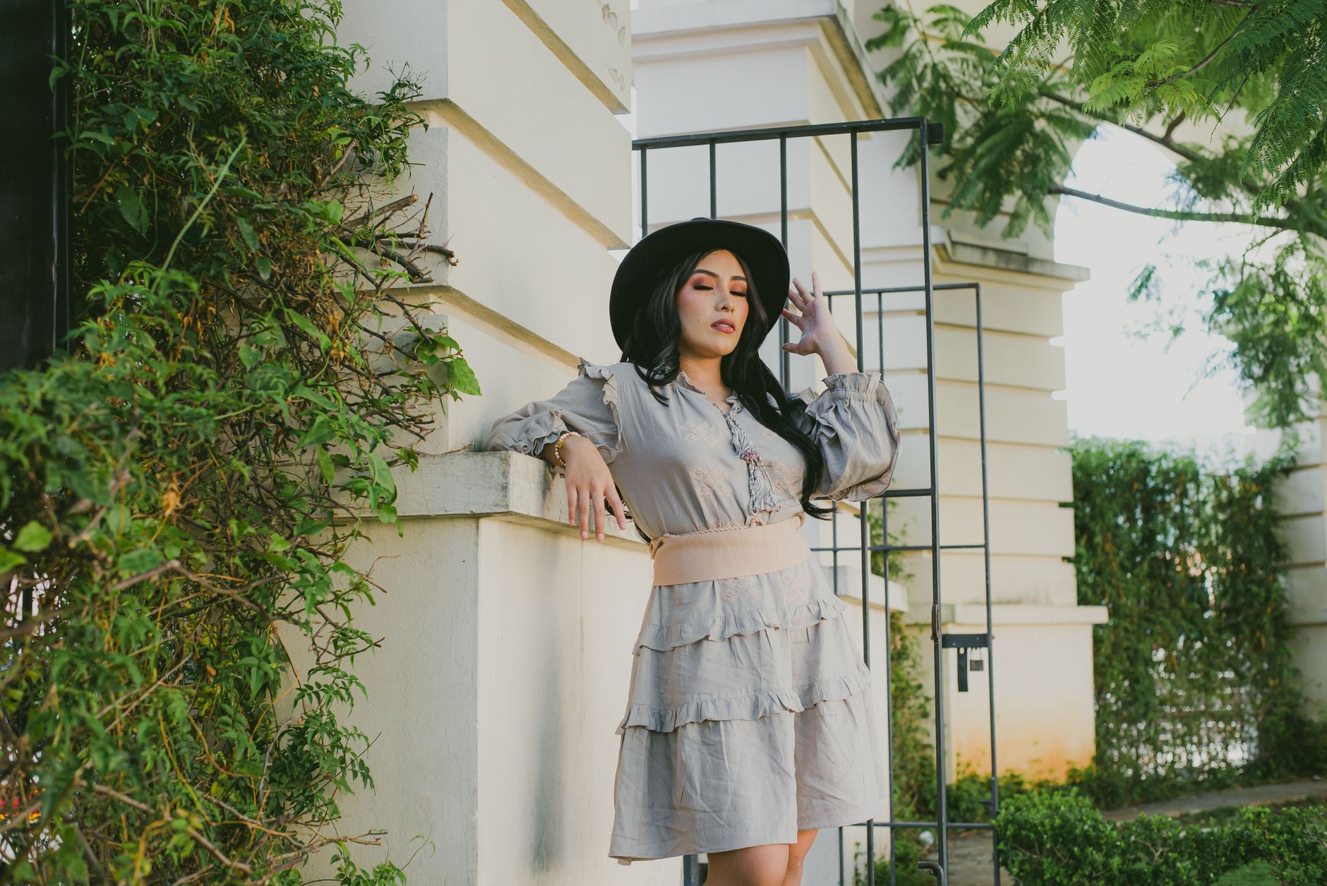 Elegant woman in a light dress and hat poses against a garden backdrop, embodying summer style.