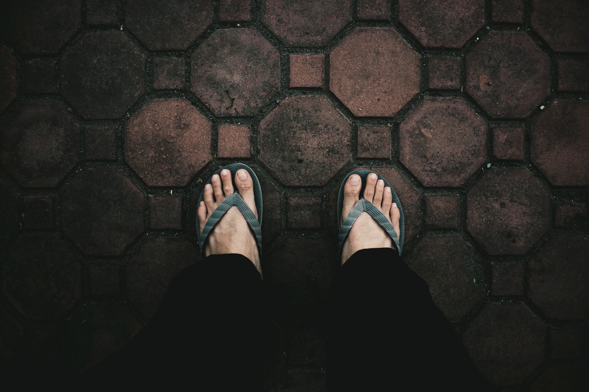 A top-down view of feet wearing flip flops standing on textured red brick pavement.