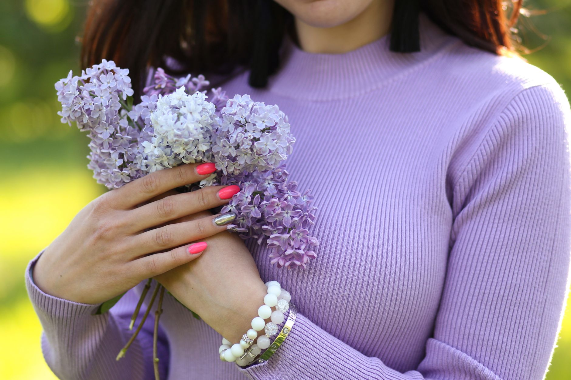 Woman's hands holding lilac flowers, wearing a lavender sweater with vibrant nails in sunlight.