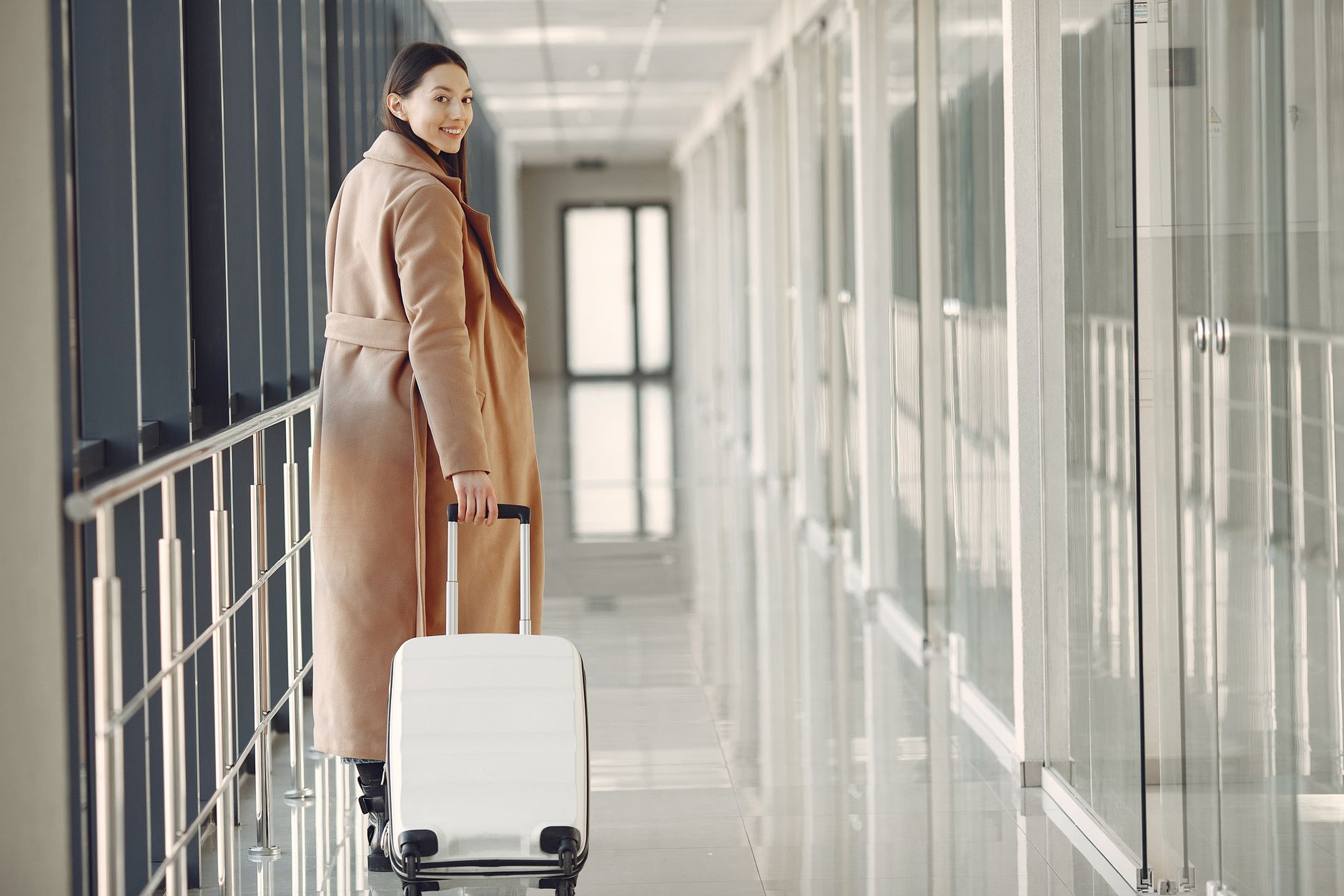 Back view full length of young cheerful female passenger in trendy beige coat walking along airport airbridge and looking at camera over sho