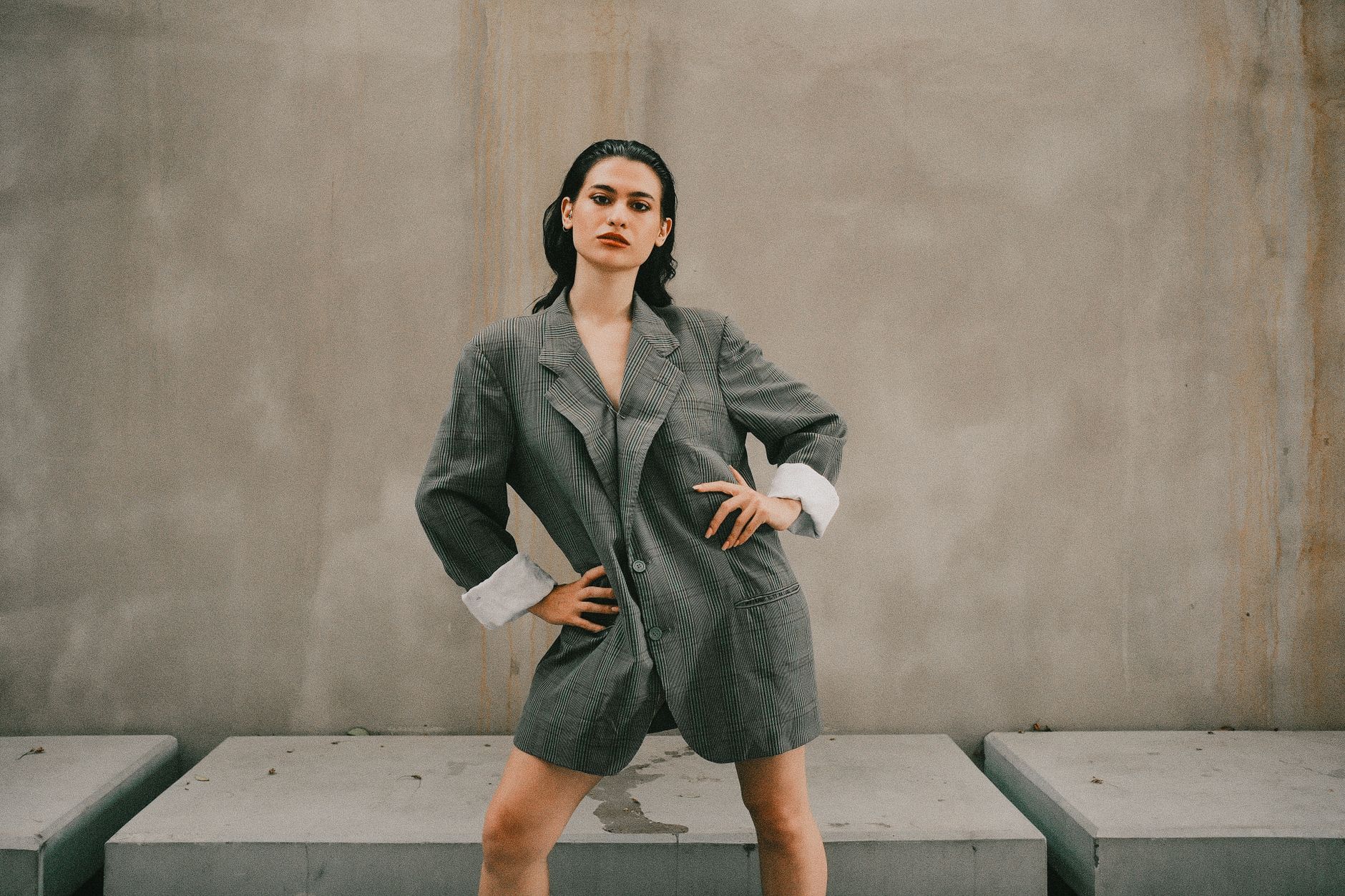 Stylish woman poses confidently in an oversized grey suit against a minimalist outdoor backdrop.