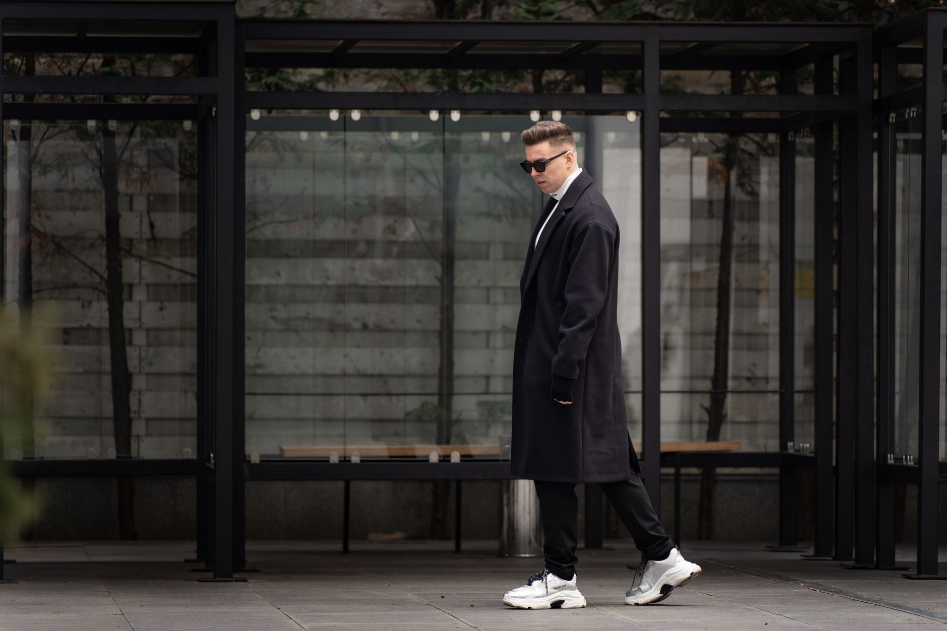 Stylish man wearing a black coat and sneakers stands confidently outdoors, exuding a modern fashion vibe.