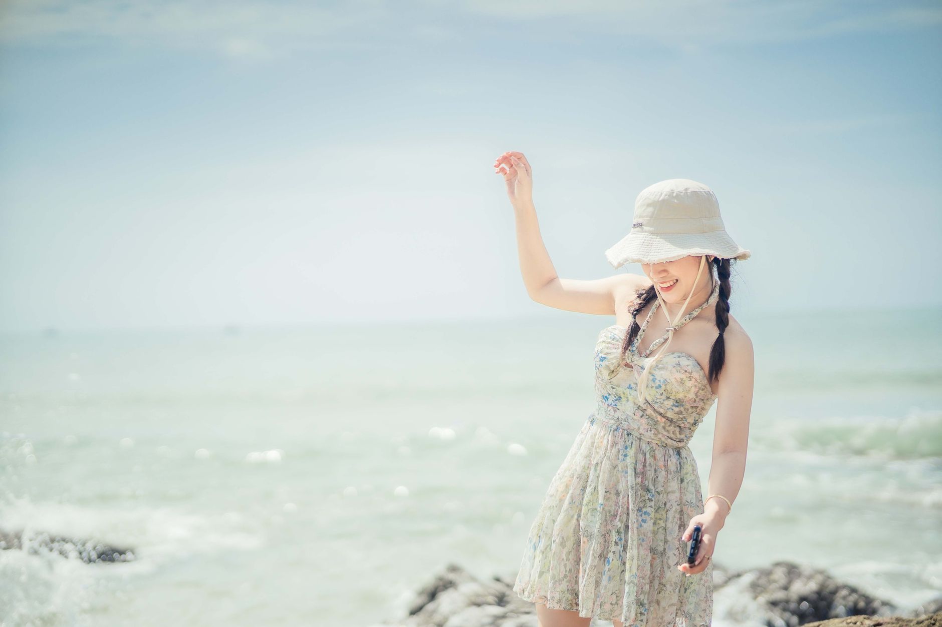 A woman in a floral sundress and bucket hat enjoys a sunny day at the beach, smiling by the sea.