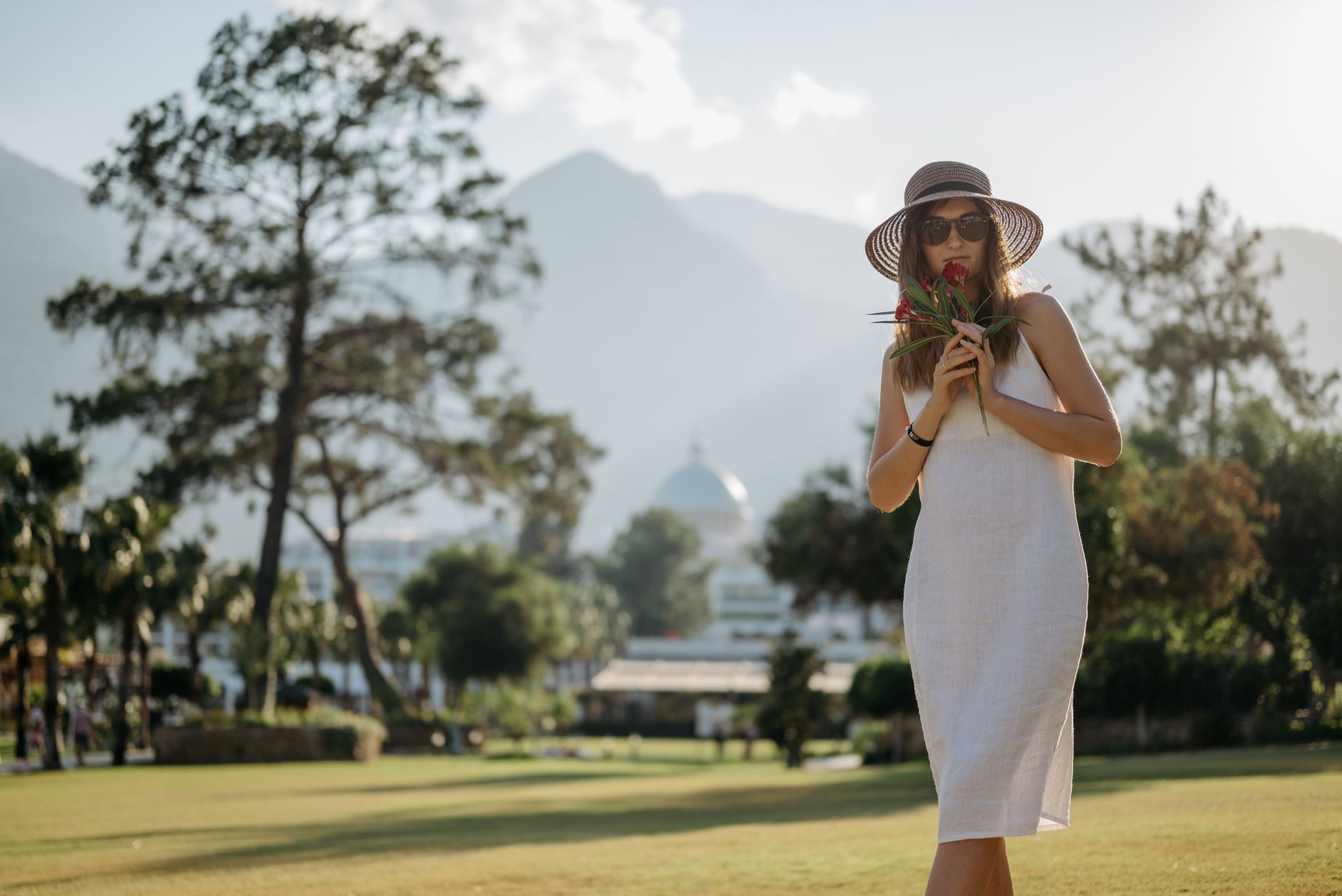 A fashionable woman in a white dress poses in a sunny park, holding a flower bouquet, wearing a straw hat and sunglasses.