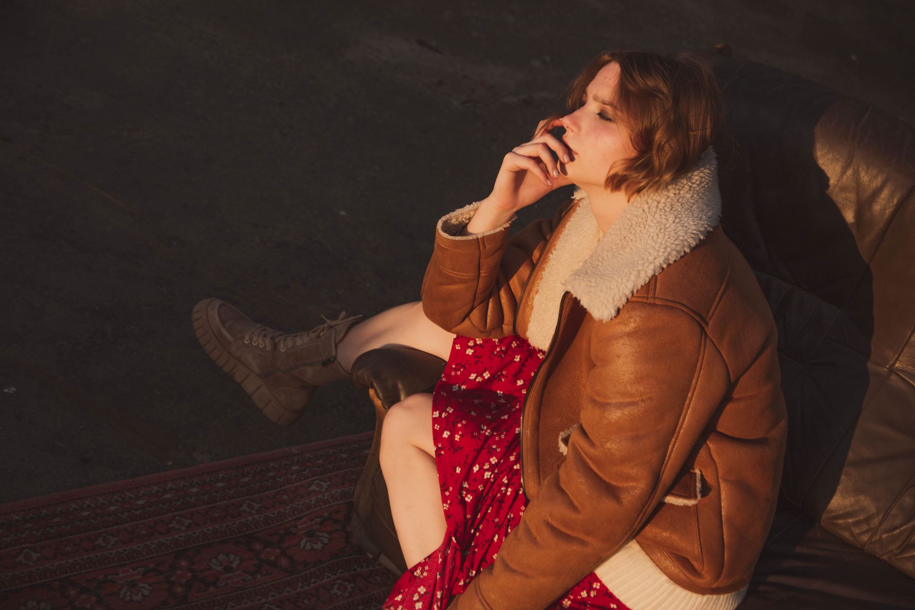 Fashionable woman in a brown leather jacket and red dress sitting contemplatively on a sofa in sunlight.