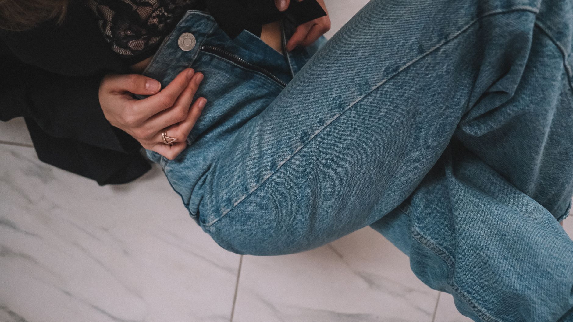 A person sitting on a marble floor, wearing denim jeans and a black jacket.