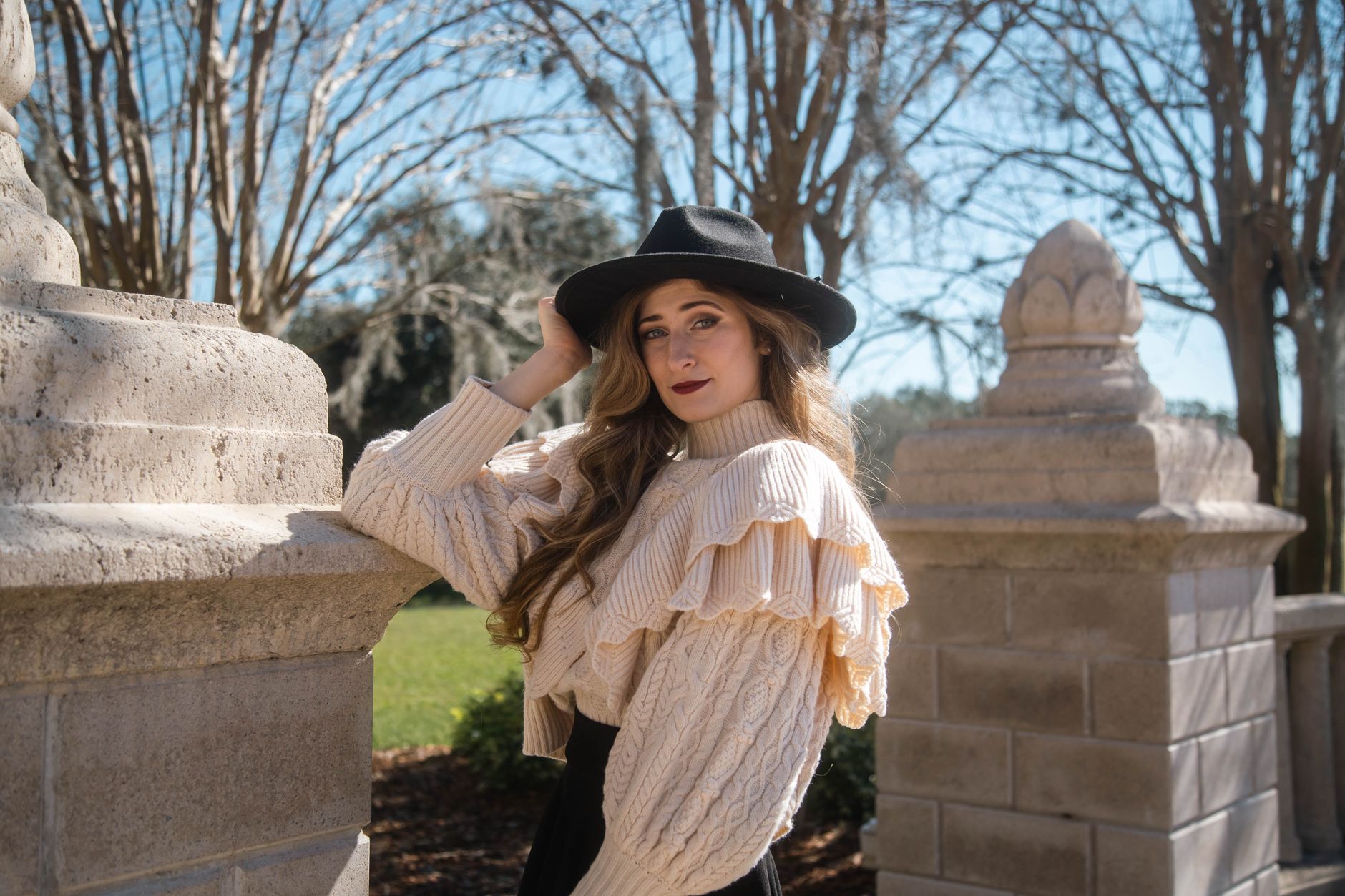 Chic woman in a ruffled cream sweater and hat posing outdoors on a sunny day.