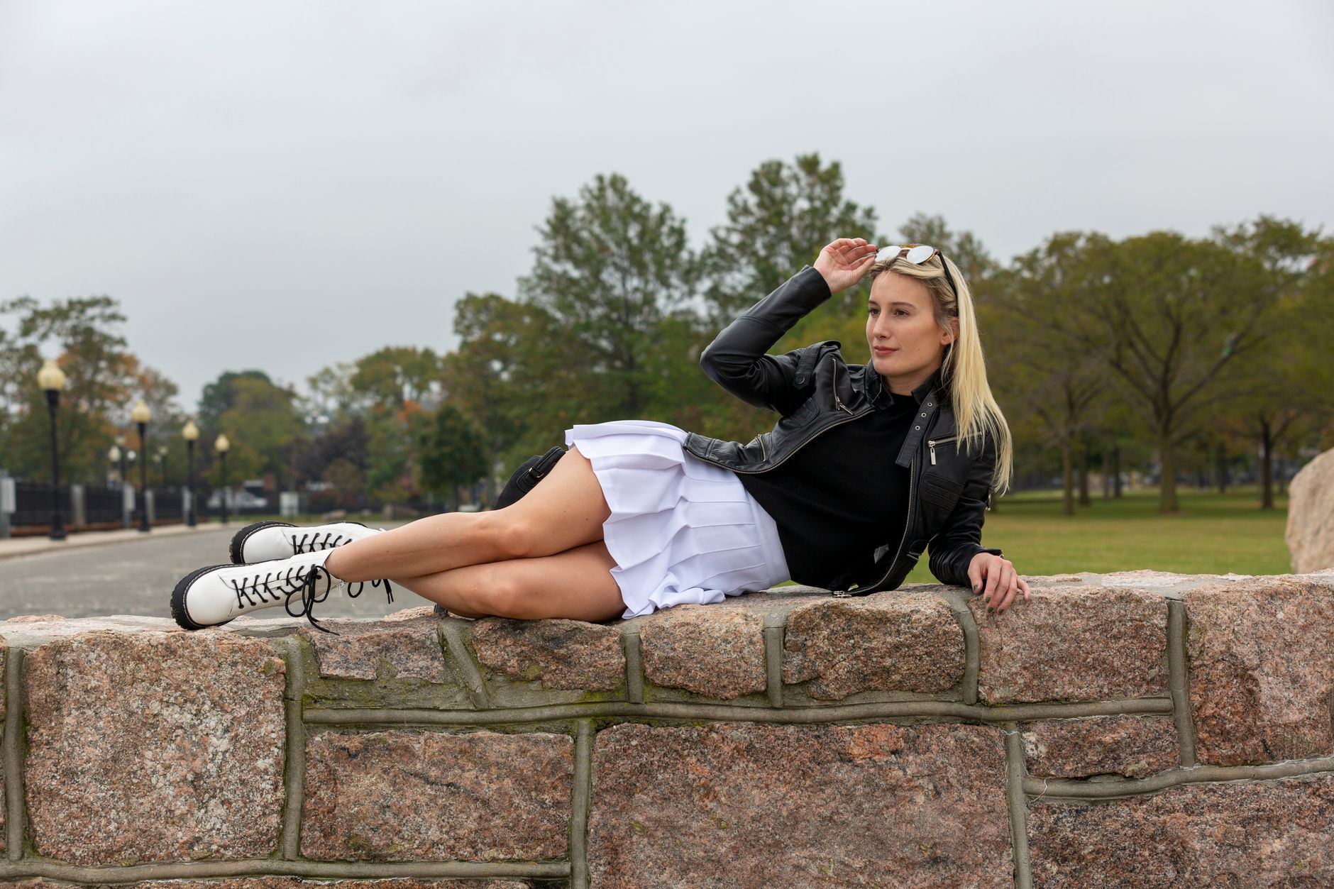 Full body of content young female with legs crossed in fashionable leather jacket and skirt resting on stone parapet