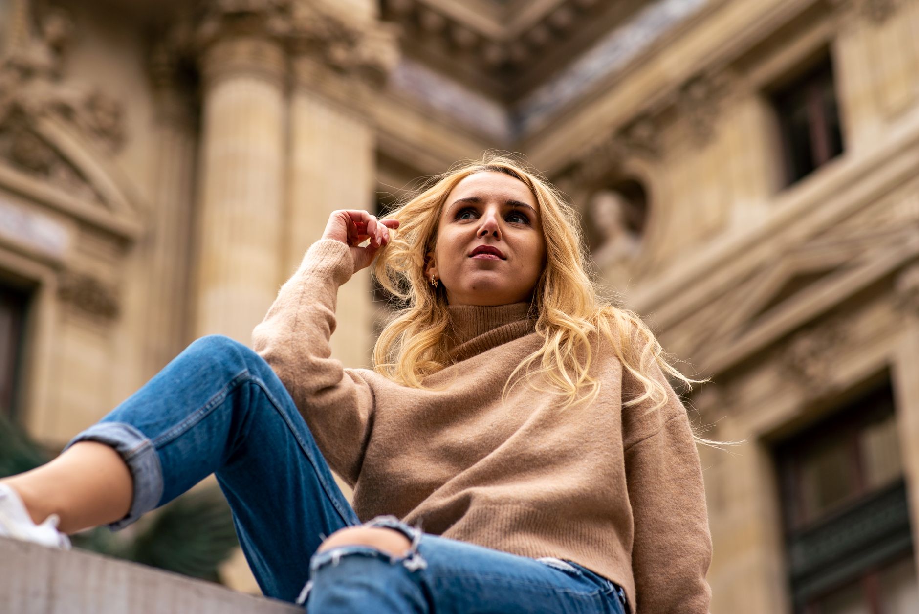 Low angle shot of a woman with blonde hair wearing a beige sweater and jeans, sitting by a historic building.
