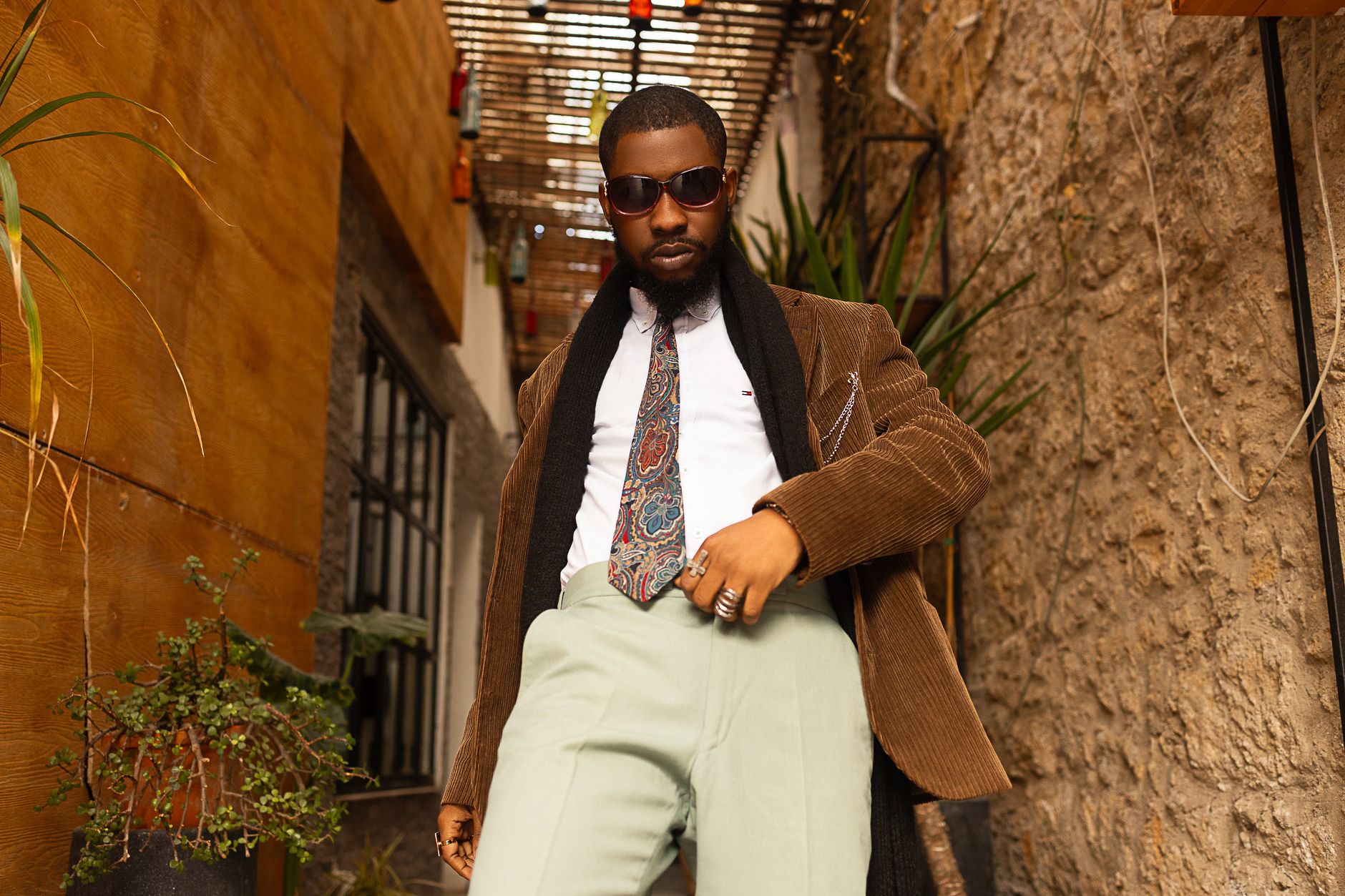 A fashionable man in a corduroy jacket poses confidently in an urban setting, wearing sunglasses and a paisley tie.