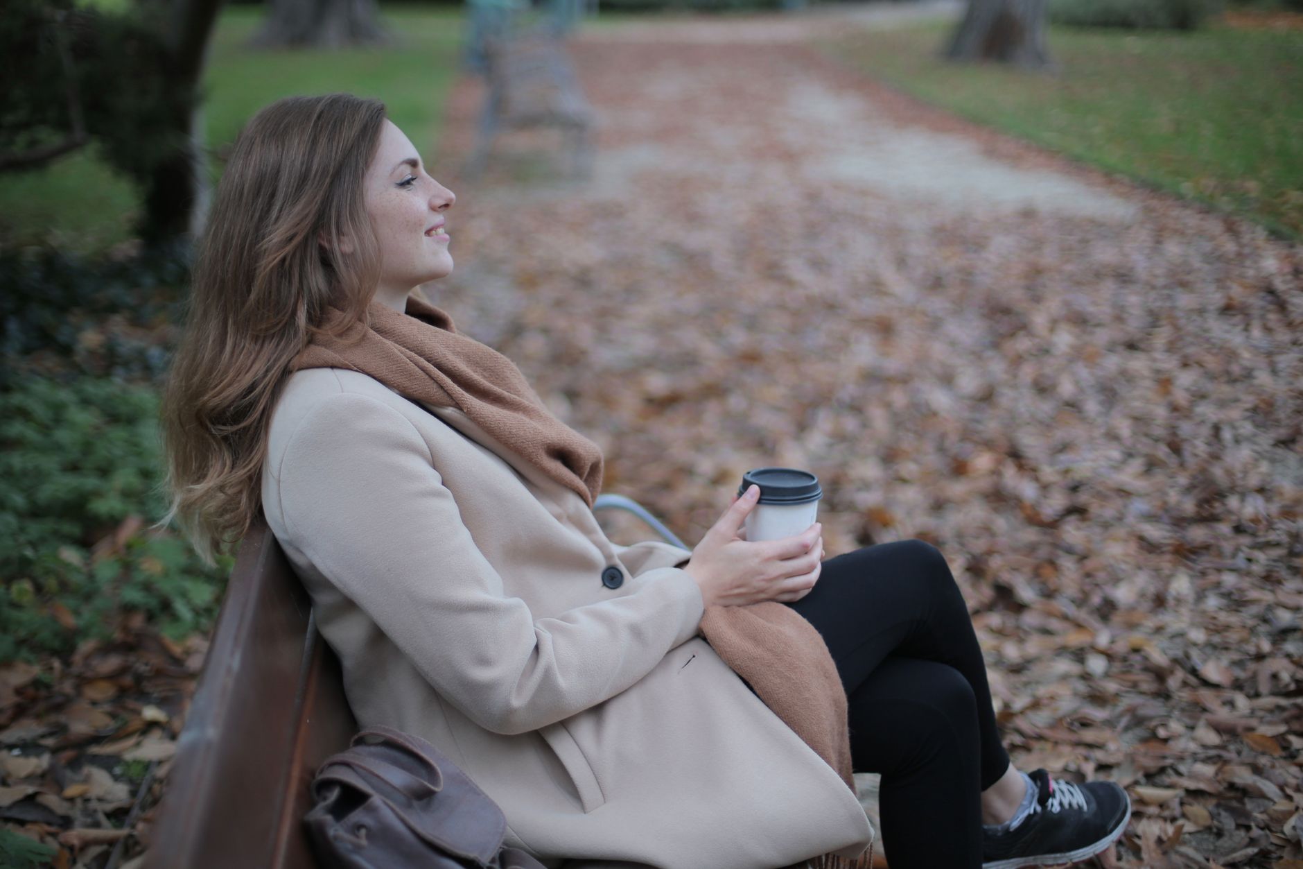 A woman in a beige coat enjoys a coffee on a park bench during autumn.