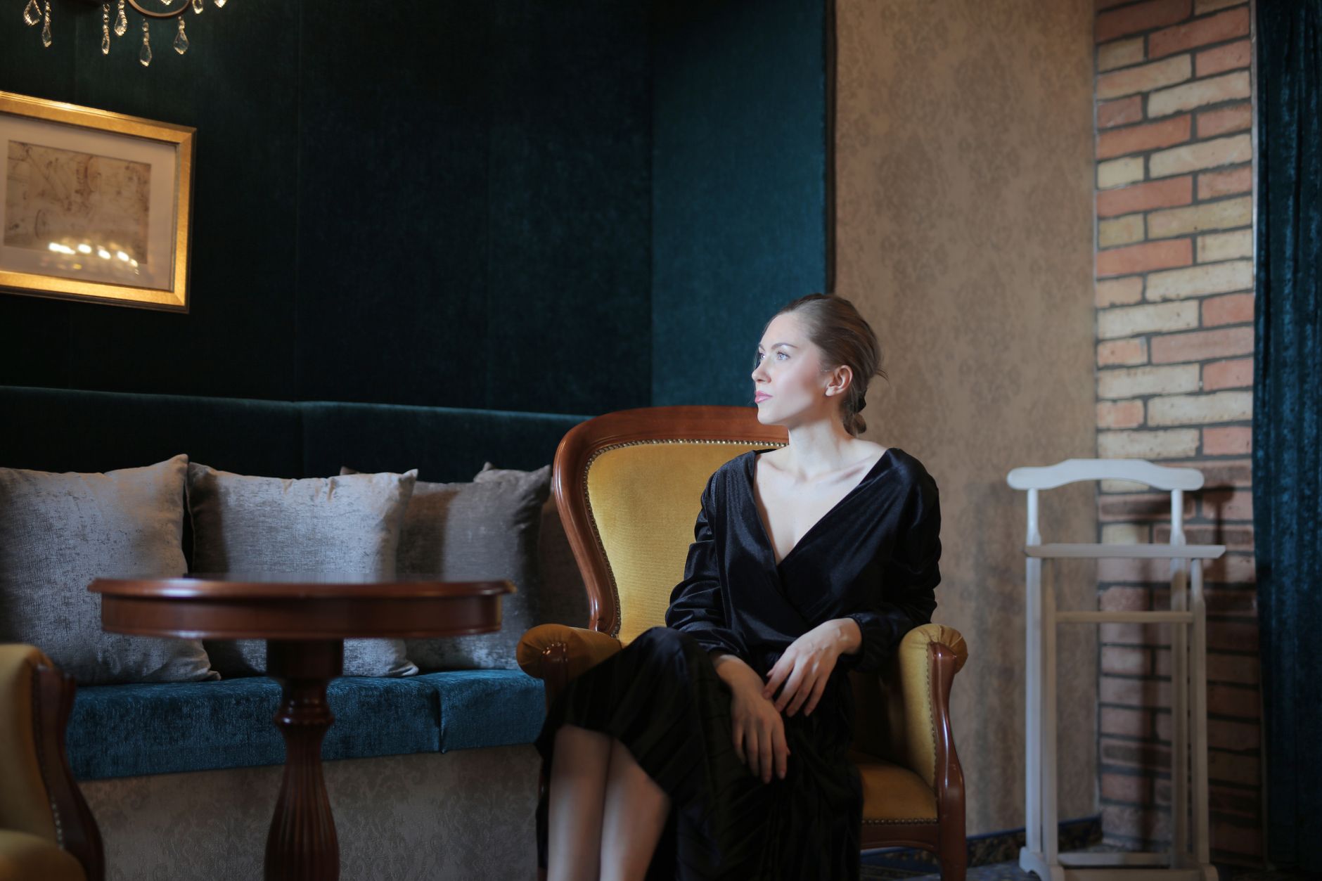 A sophisticated woman in a black dress sits in a vintage armchair in a lush, cozy interior.