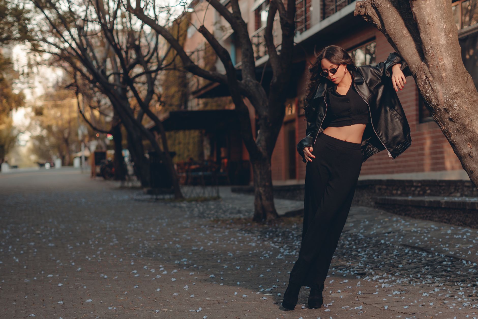 Fashionable woman in a black leather jacket posing stylishly on a tree-lined urban street.