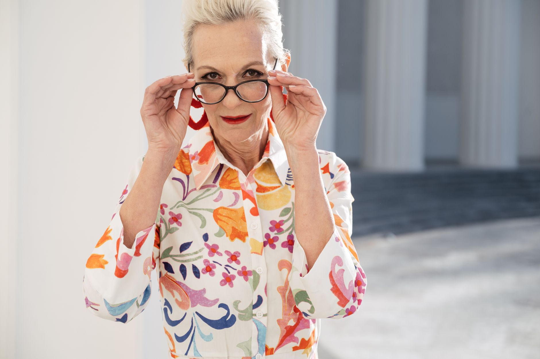 Confident senior woman in floral dress adjusting glasses outdoors on a sunny day.