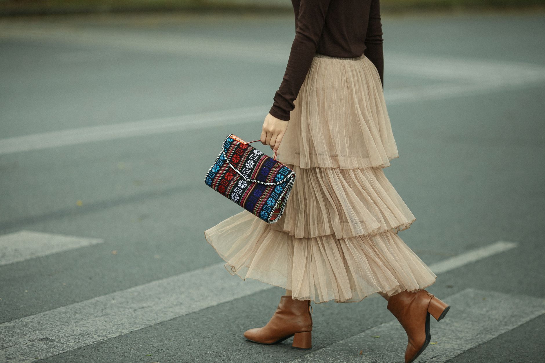 Woman in elegant skirt and boots crossing a zebra crossing, holding a boho-style bag.