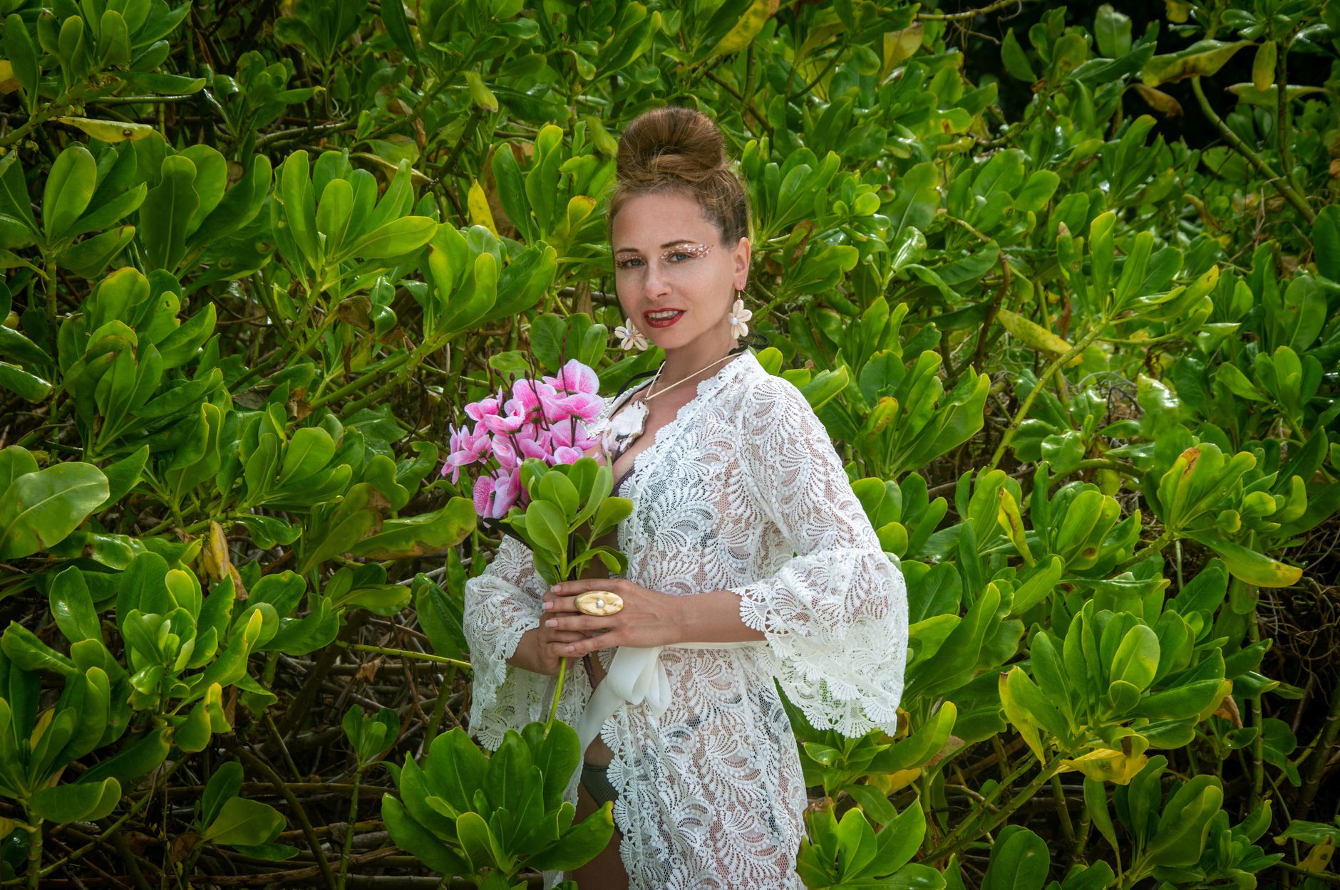 Elegant woman in a lace dress holding pink flowers, standing amidst lush green foliage.