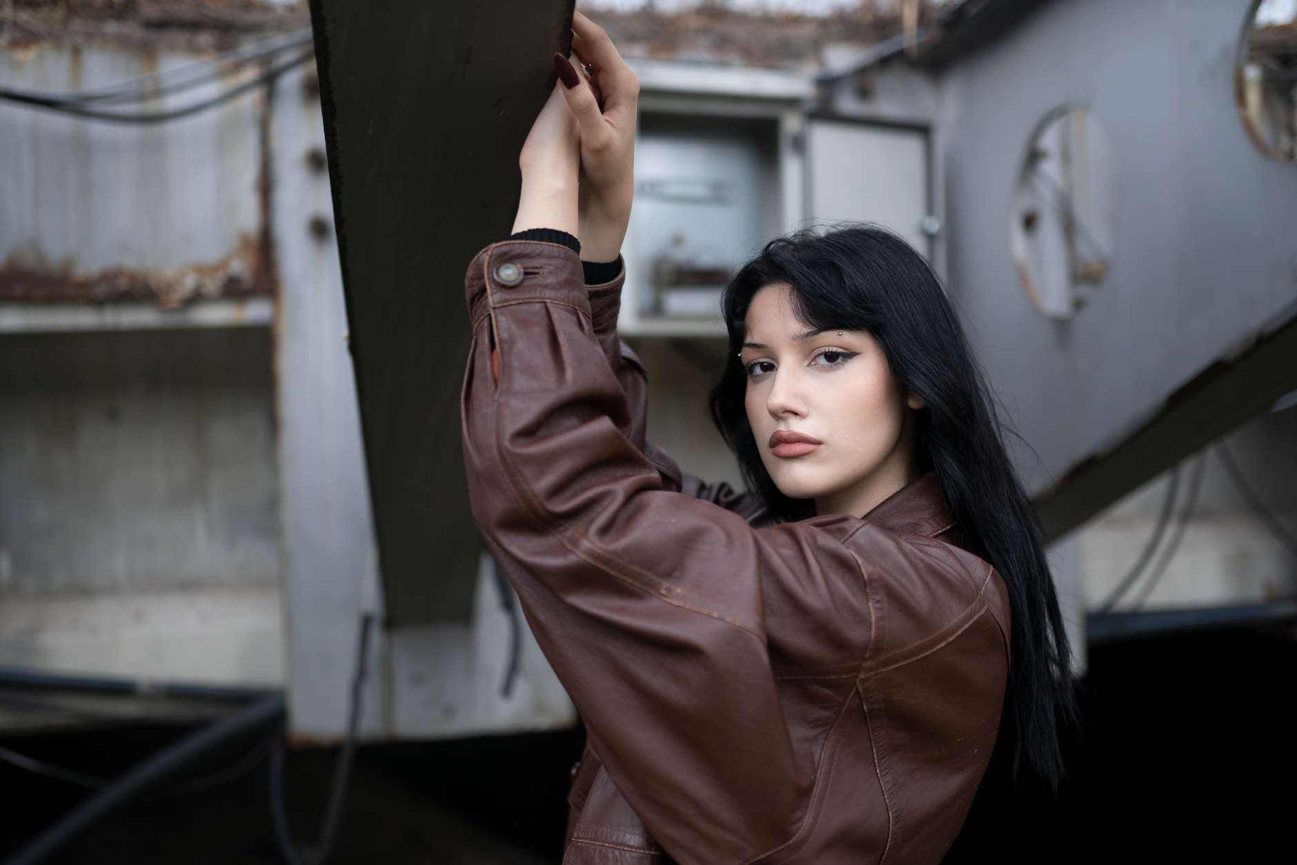 Young woman posing with a confident expression in a brown leather jacket in an abandoned industrial area.