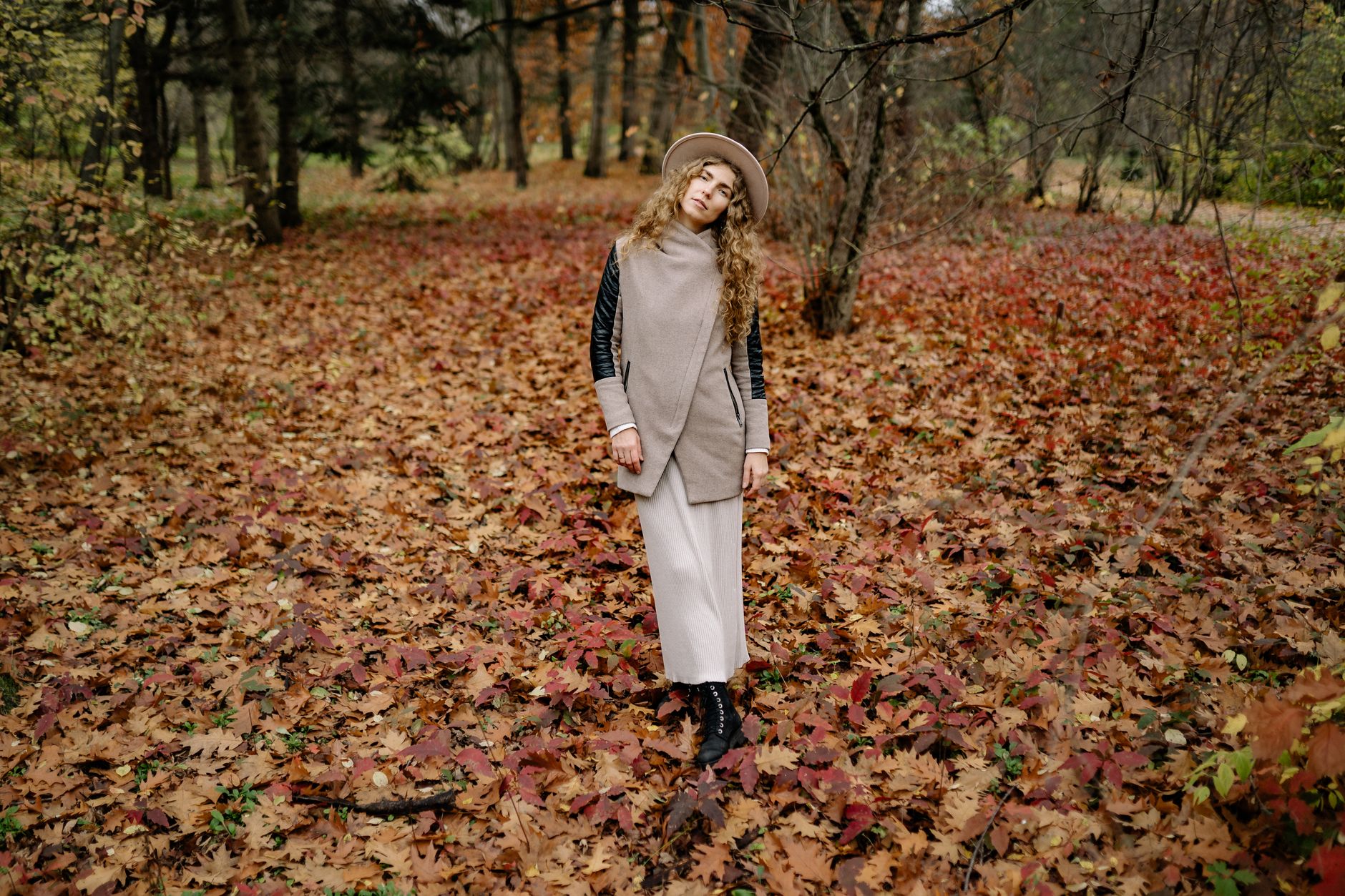Stylish woman in warm clothing poses amidst colorful autumn leaves in a serene forest setting.