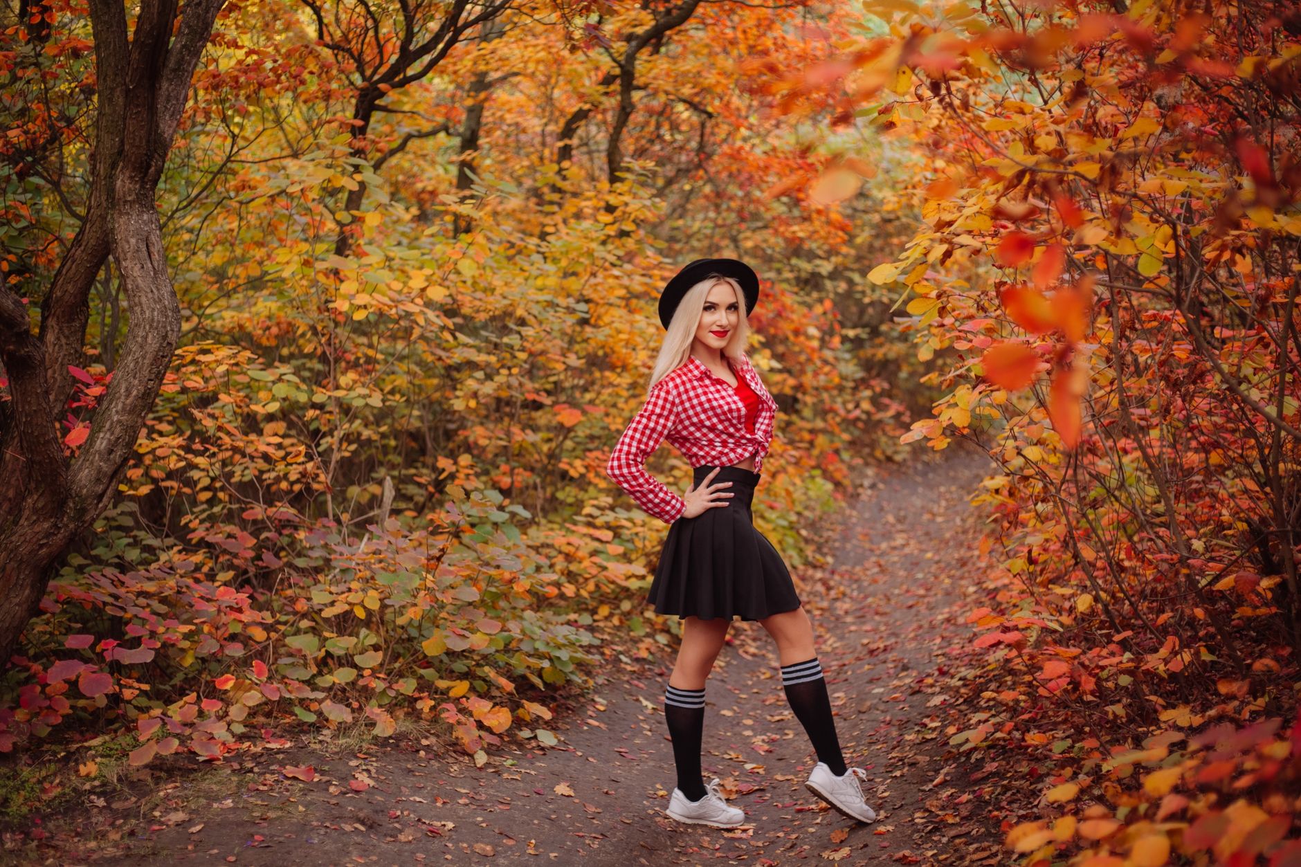 A stylish woman poses in a colorful autumn forest, surrounded by vibrant leaves.