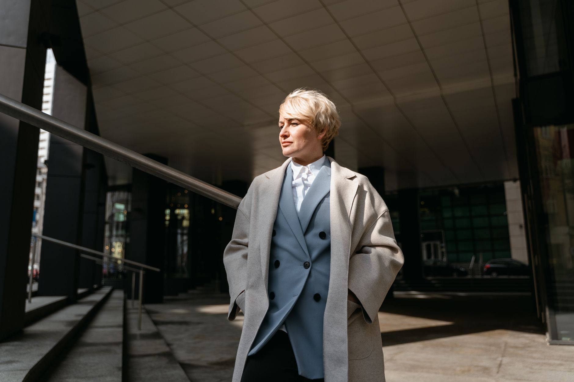 Confident blonde woman in a stylish overcoat, standing in an urban setting with hands in pockets.