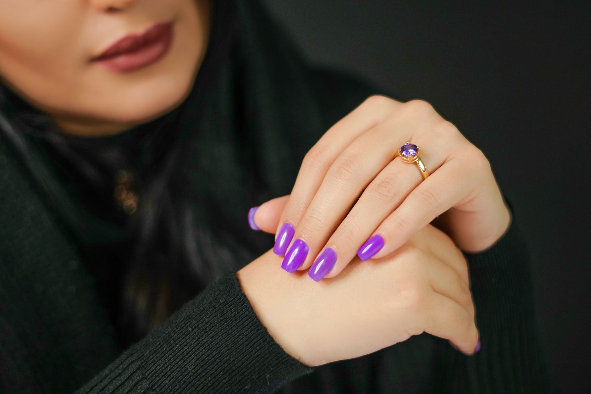 Close-up of woman's hands with vibrant purple nails and a gold ring, set against a dark background.
