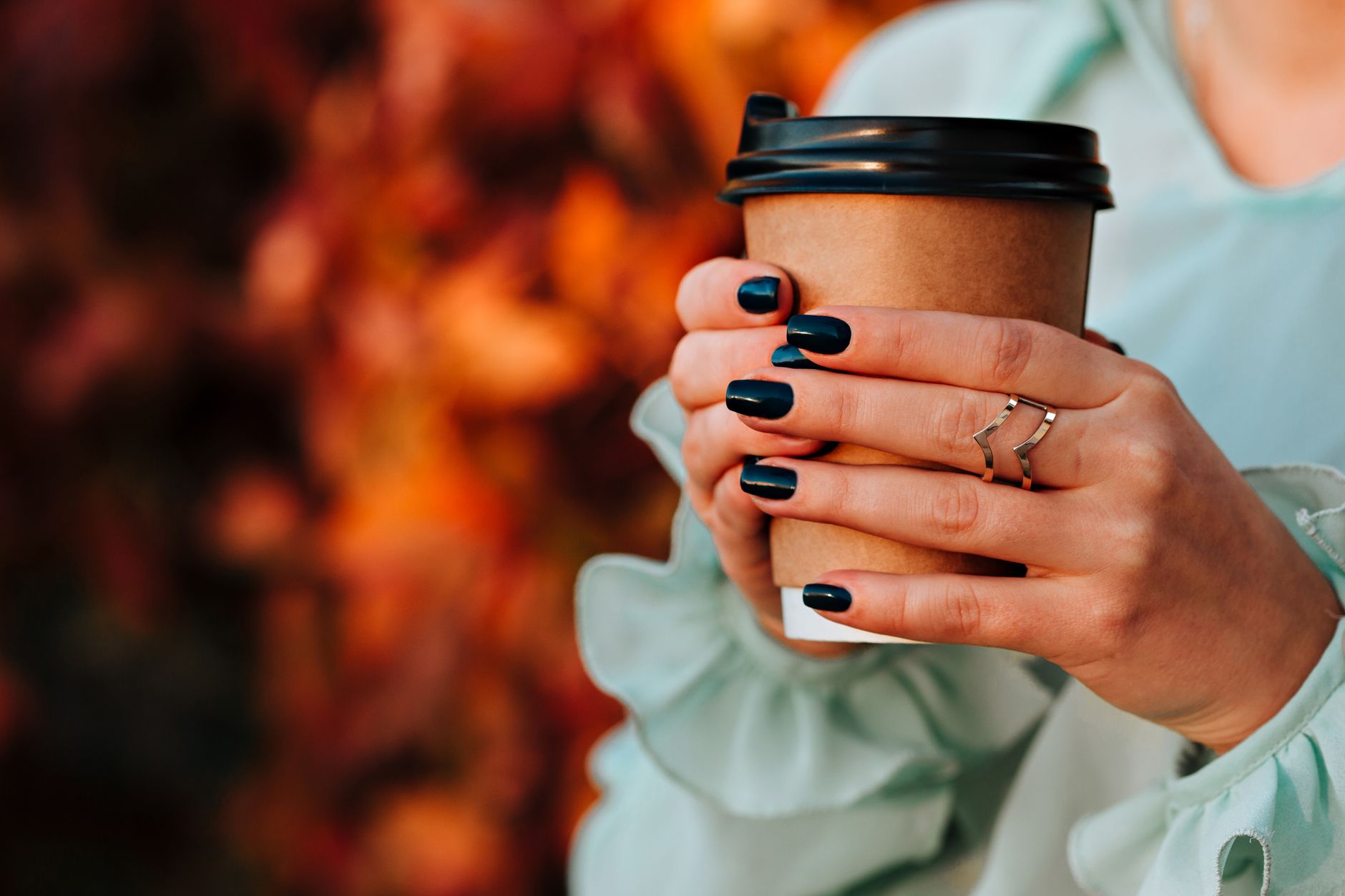 A woman's hands with dark nail polish holding a paper coffee cup against an autumn backdrop.