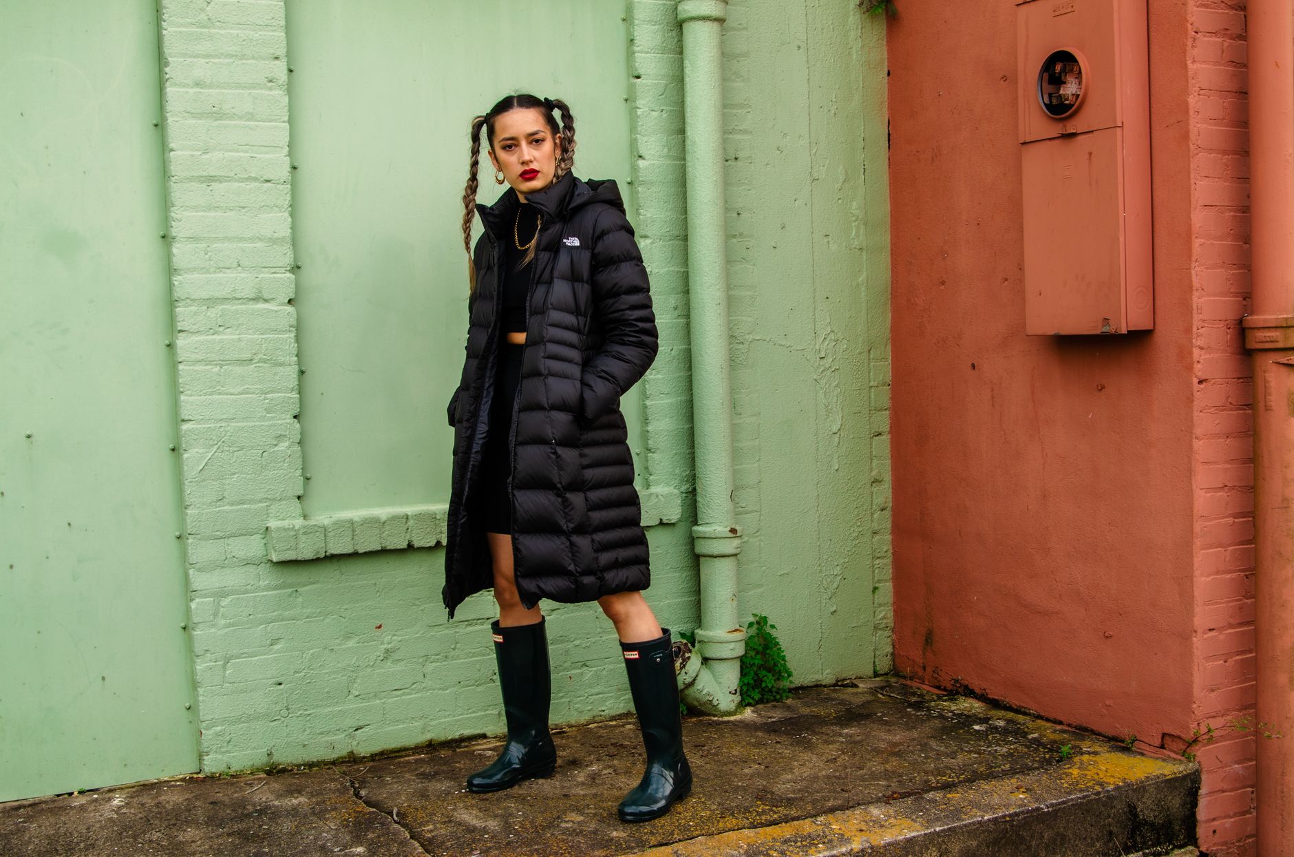Fashionable woman in black coat and rain boots standing against a green and terracotta wall outdoors.
