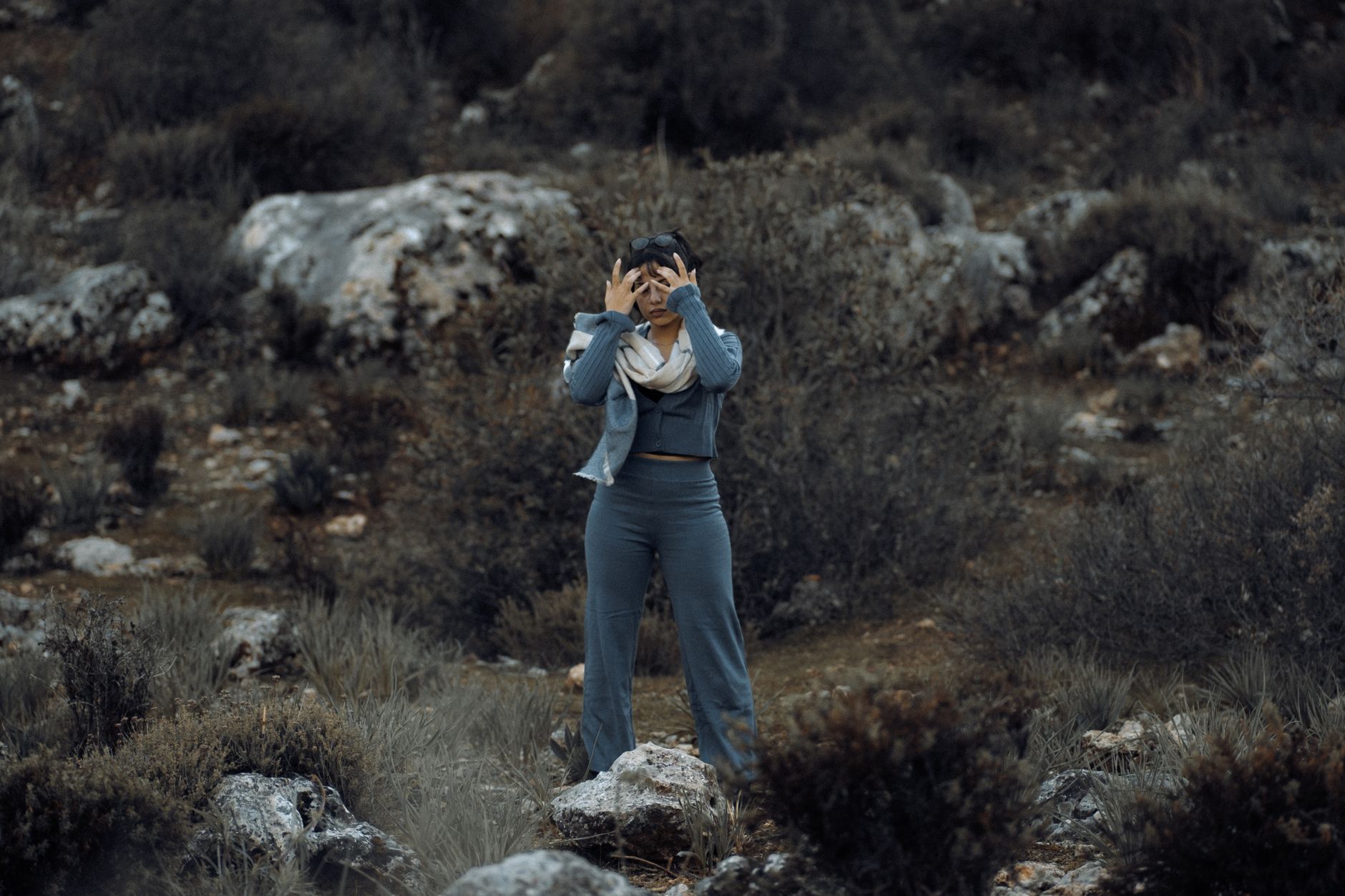 Fashionable woman stands in an outdoor rocky terrain wearing gray attire and a scarf.