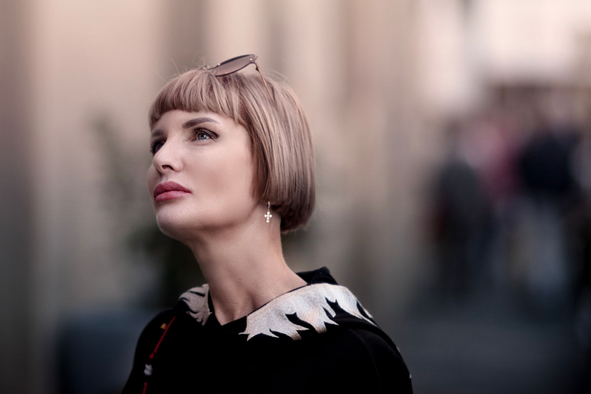 Stylish portrait of a woman with blonde bob haircut and sunglasses, looking up with a blurred urban background.