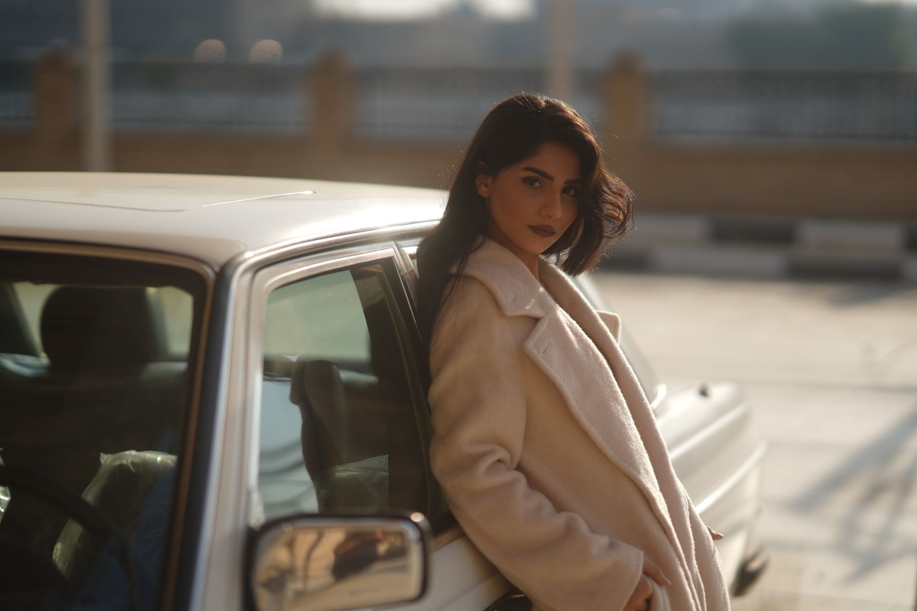 Stylish woman in a beige coat leaning on a vintage car in warm sunlight, creating a nostalgic outdoor scene.