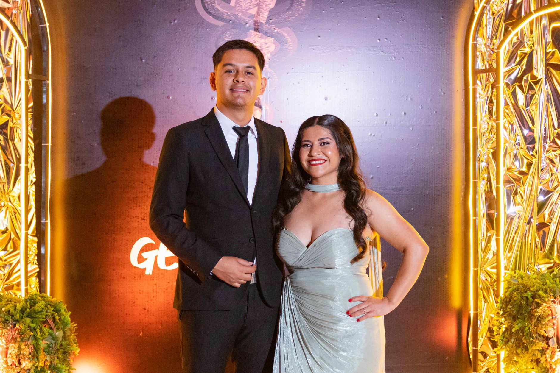 Stylish couple in formal attire smiling at a glamorous event with golden decor.