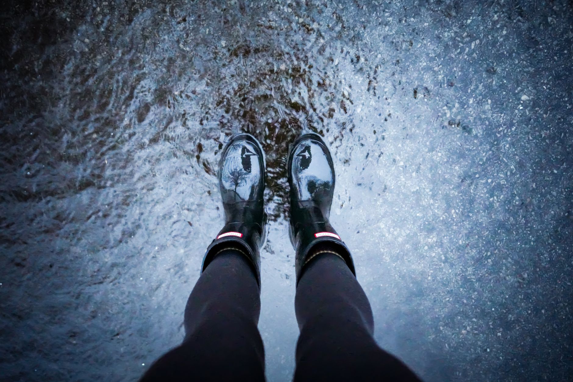 A top-down view of a person in black rain boots standing in a puddle, creating reflections.