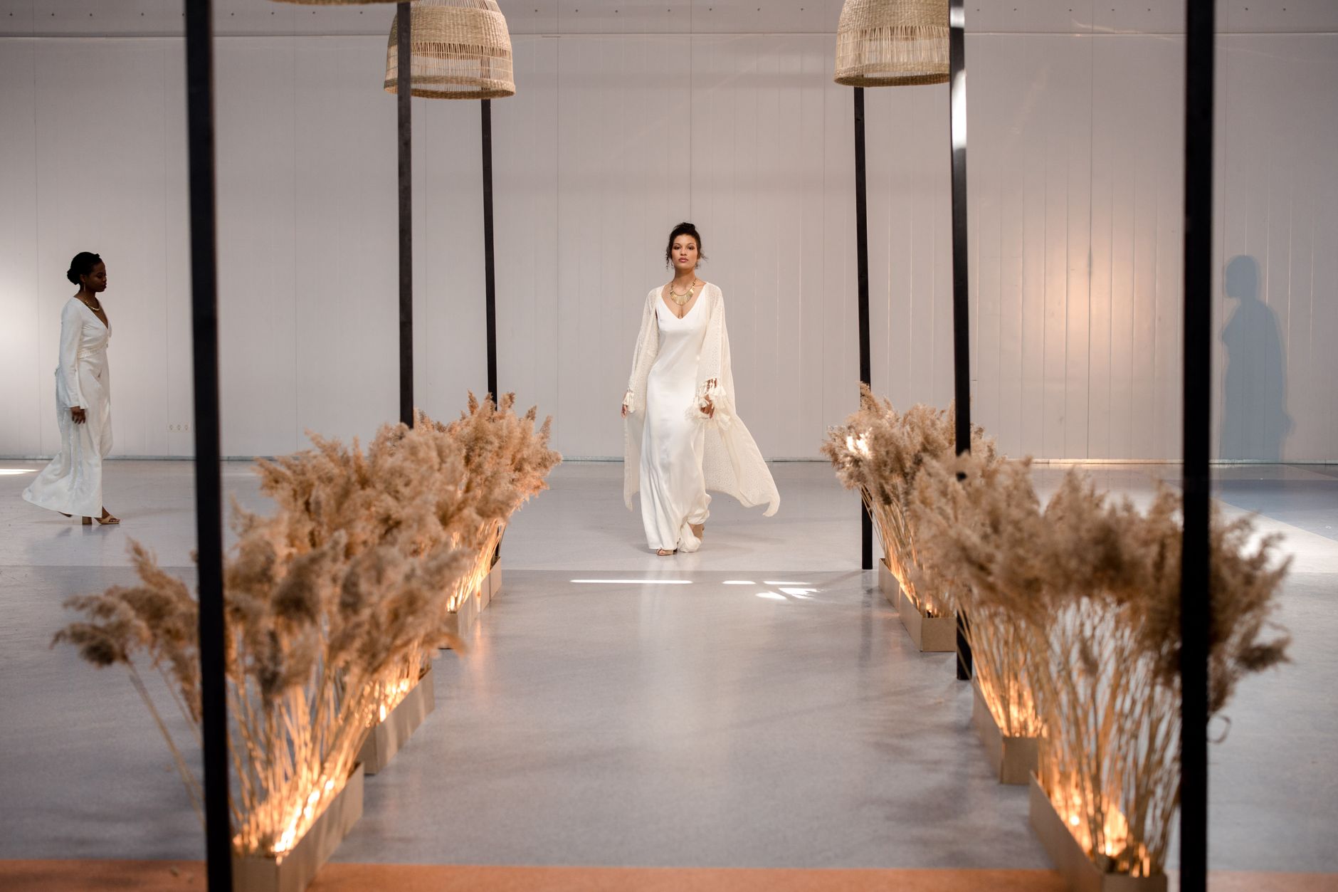 Models walking down a stylish runway in white dresses during an elegant indoor fashion show.