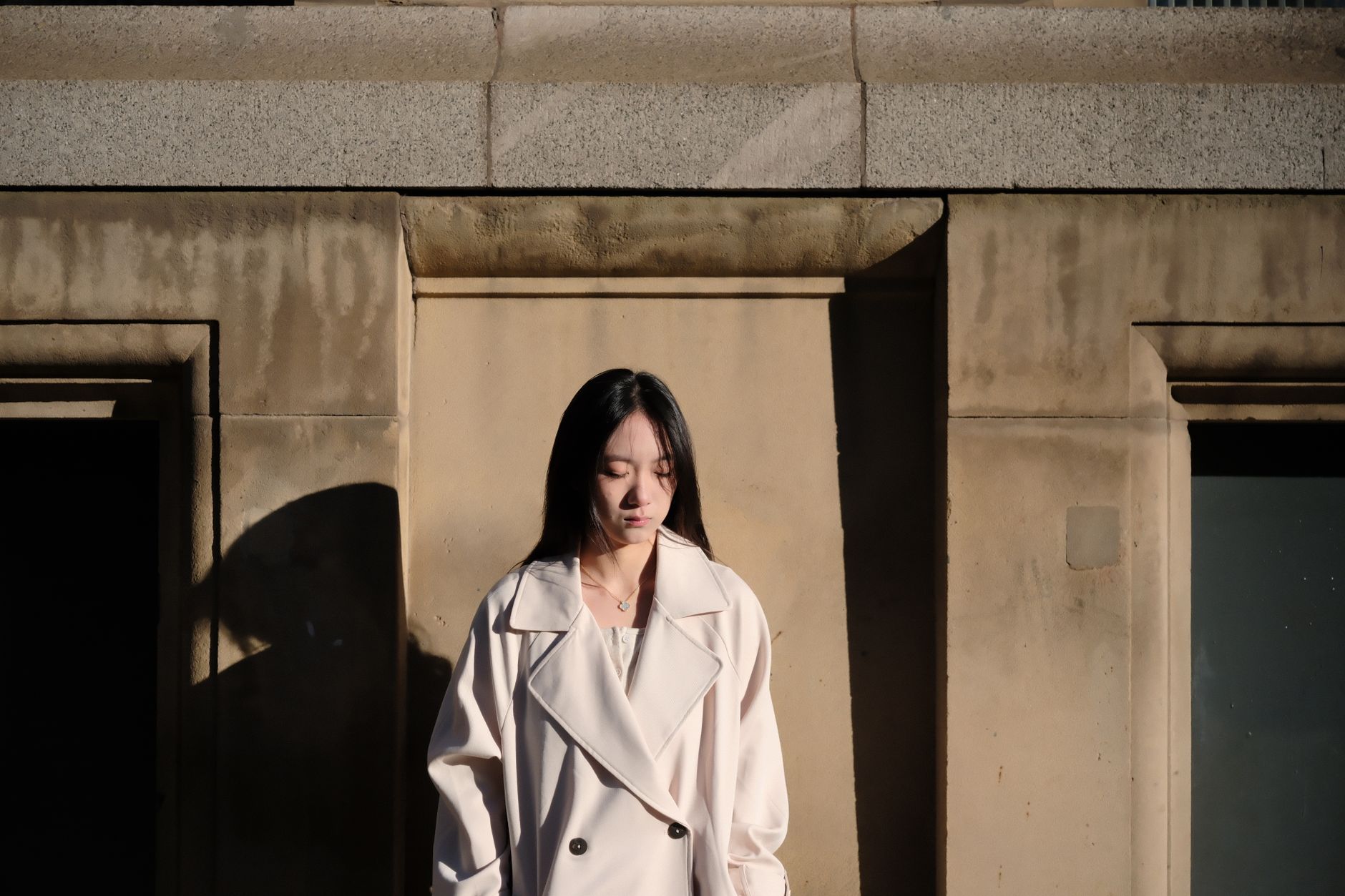 A woman in a cream trench coat stands against a stone wall, bathed in sunlight.
